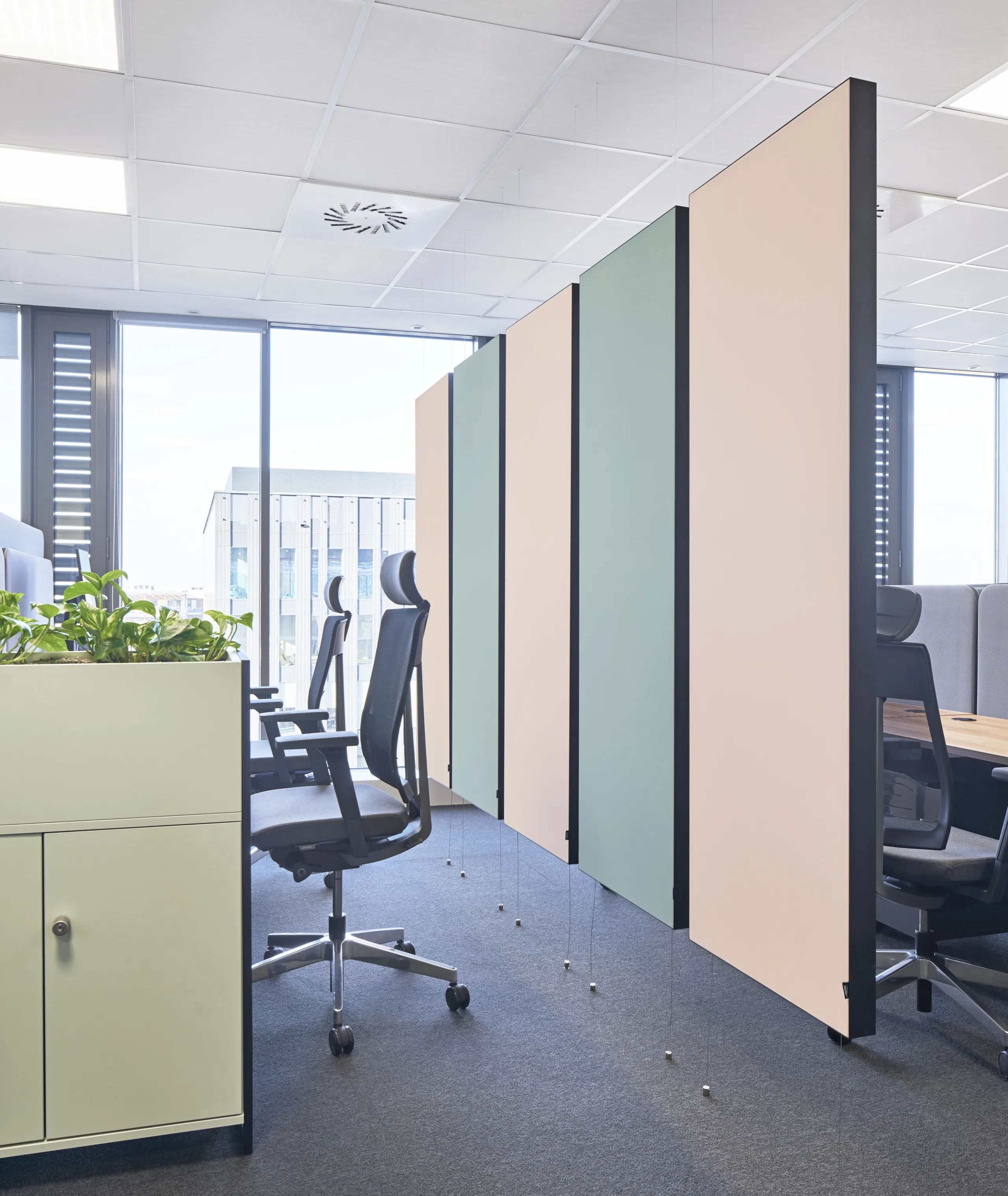 Modern office space with suspended pastel acoustic panels, workstation pods, plant-filled modular partitions, and a white grid acoustic ceiling.