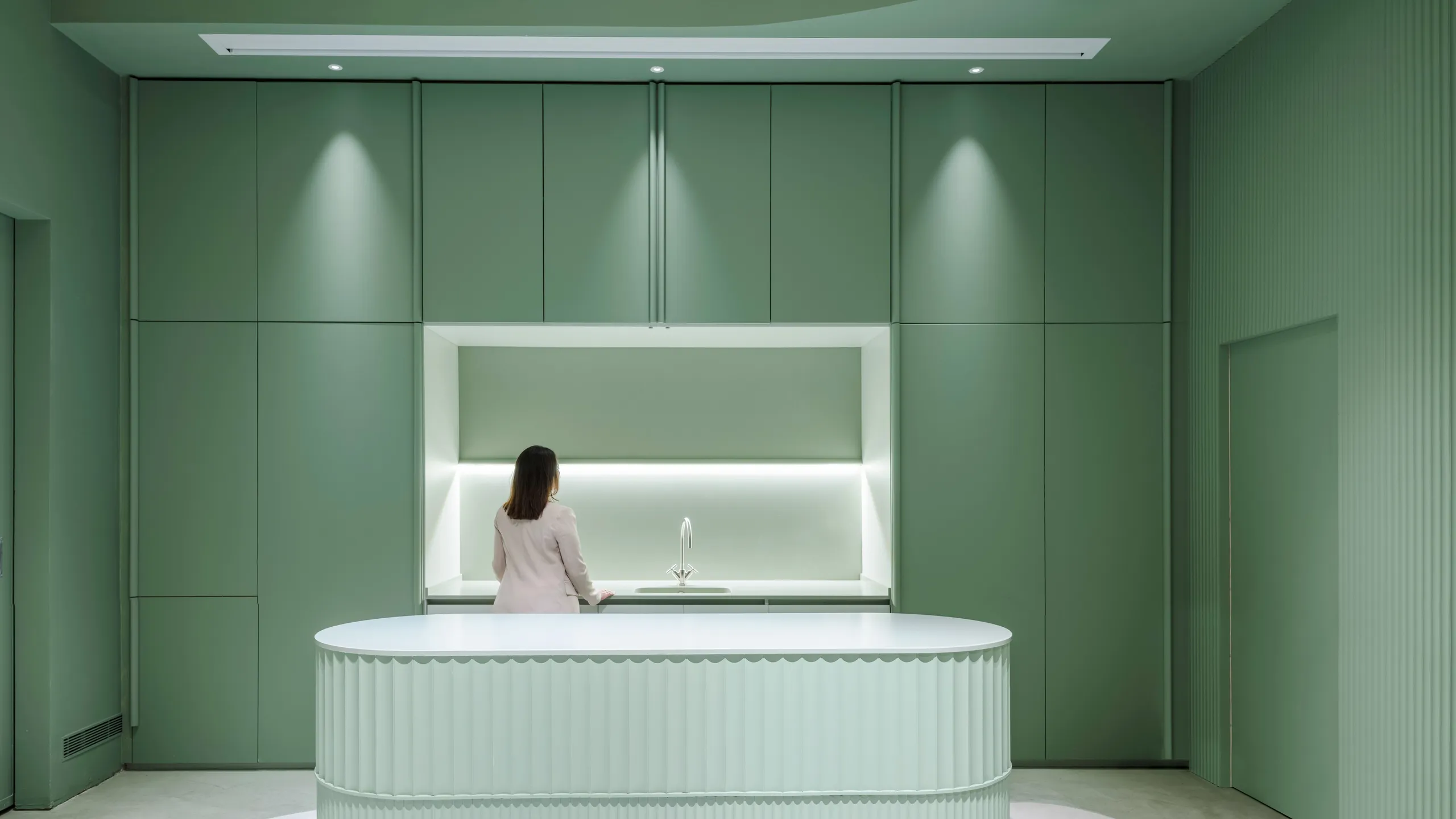 Minimalistic green kitchen with a curved island, sleek cabinetry, and modern lighting. A person stands at the countertop.