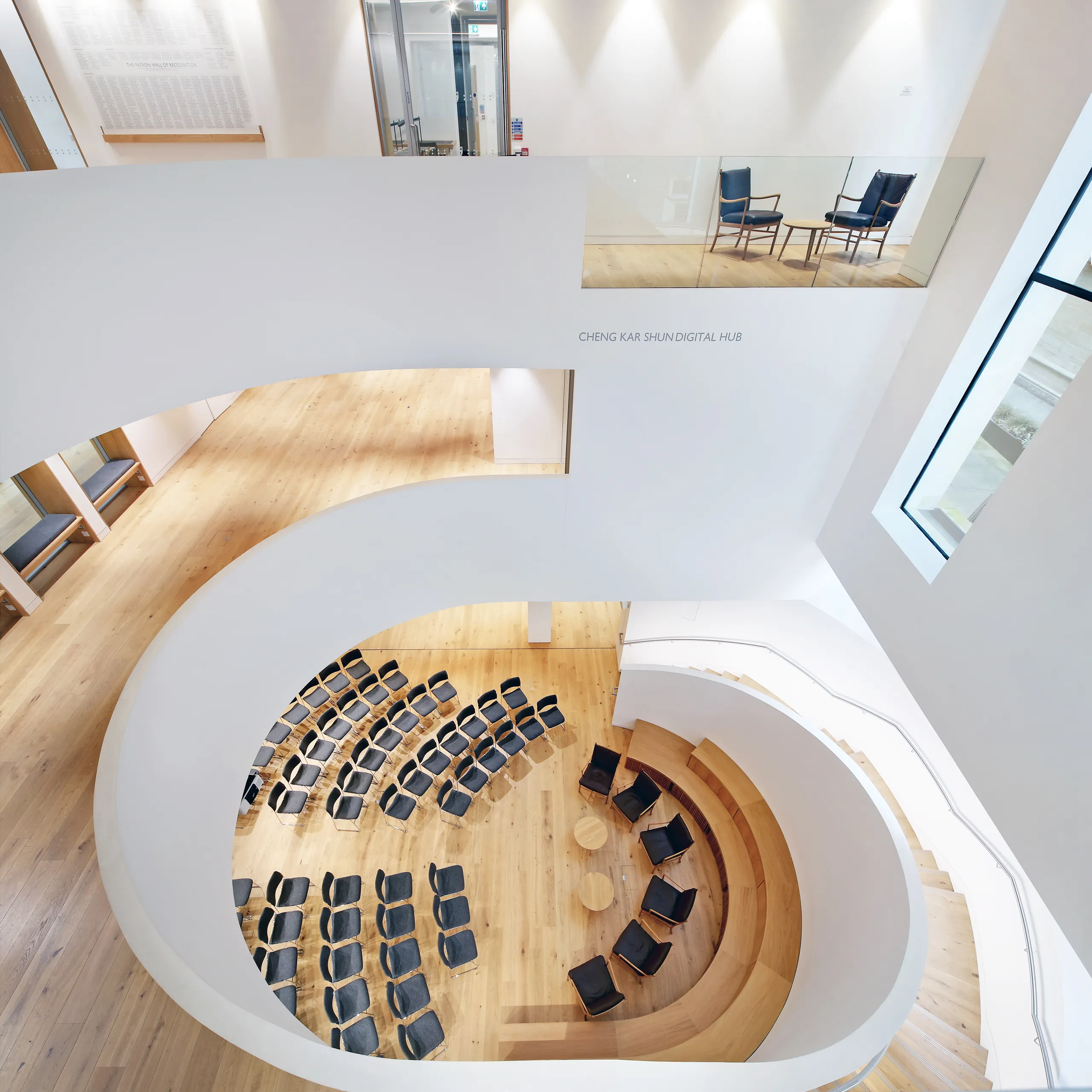 A light-filled atrium featuring curved white acoustic ceilings, a circular tiered seating area with wooden steps, and rows of black chairs arranged for presentations.