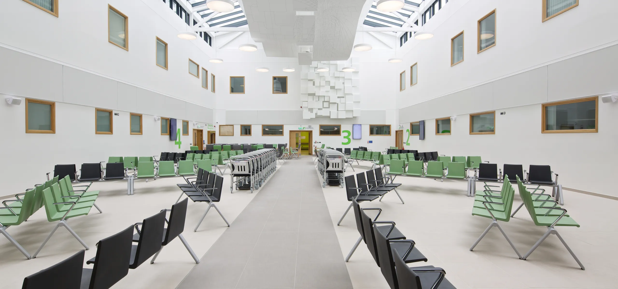 Bright healthcare atrium featuring bespoke white acoustic islands with geometric detailing, suspended under a full-height skylight to control noise in the busy waiting area.