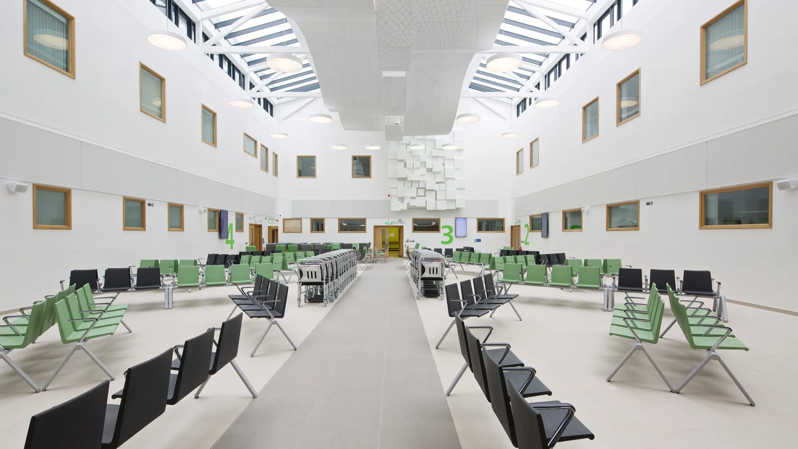 Spacious atrium with a floating raft ceiling and skylights, green and black seating areas, and geometric ceiling installations; modern and bright ambiance.