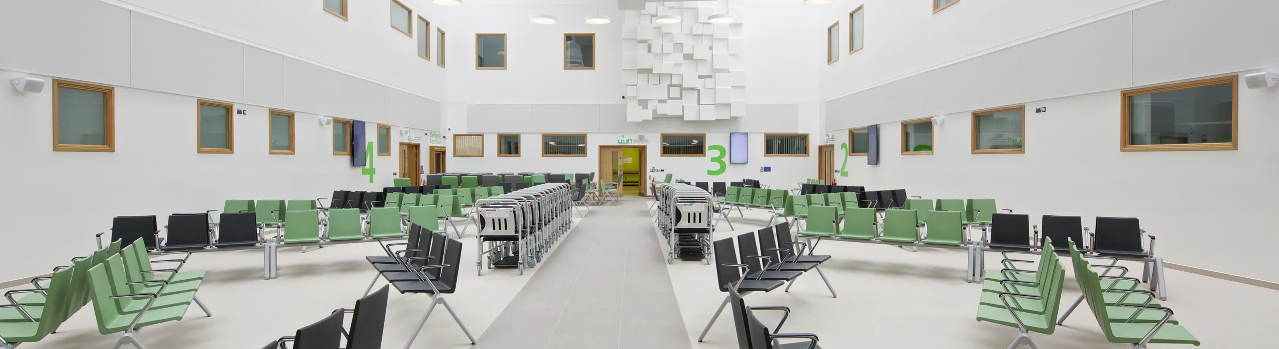 Bright healthcare atrium featuring bespoke white acoustic islands with geometric detailing, suspended under a full-height skylight to control noise in the busy waiting area.