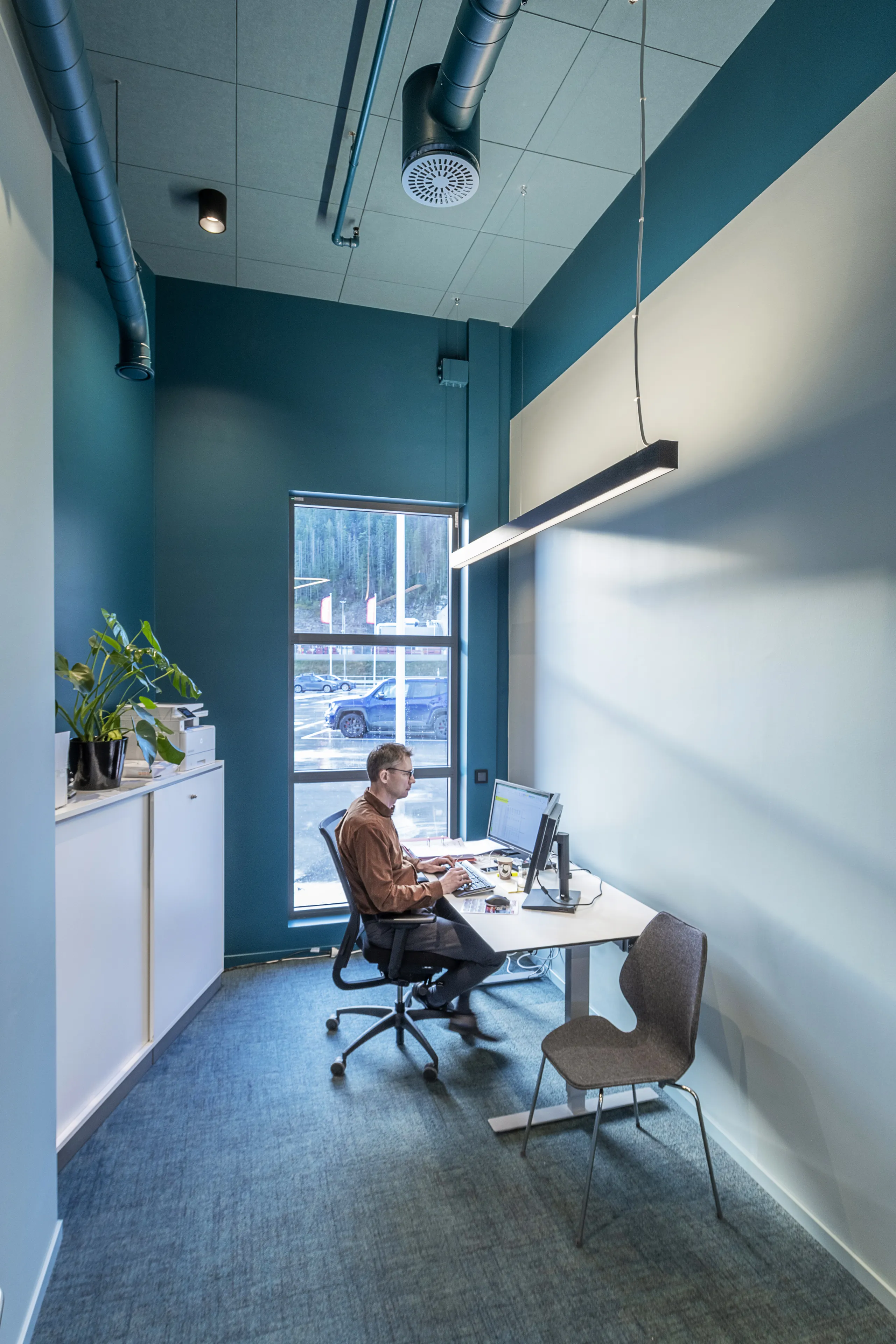 Contemporary single-person office featuring teal accent walls, high ceiling with exposed ducts, large window, and minimalist workstation setup