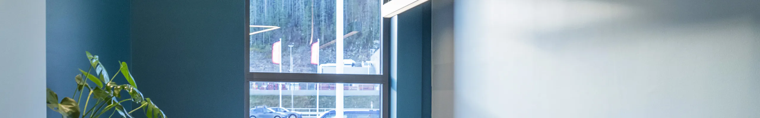 Contemporary single-person office featuring teal accent walls, high ceiling with exposed ducts, large window, and minimalist workstation setup