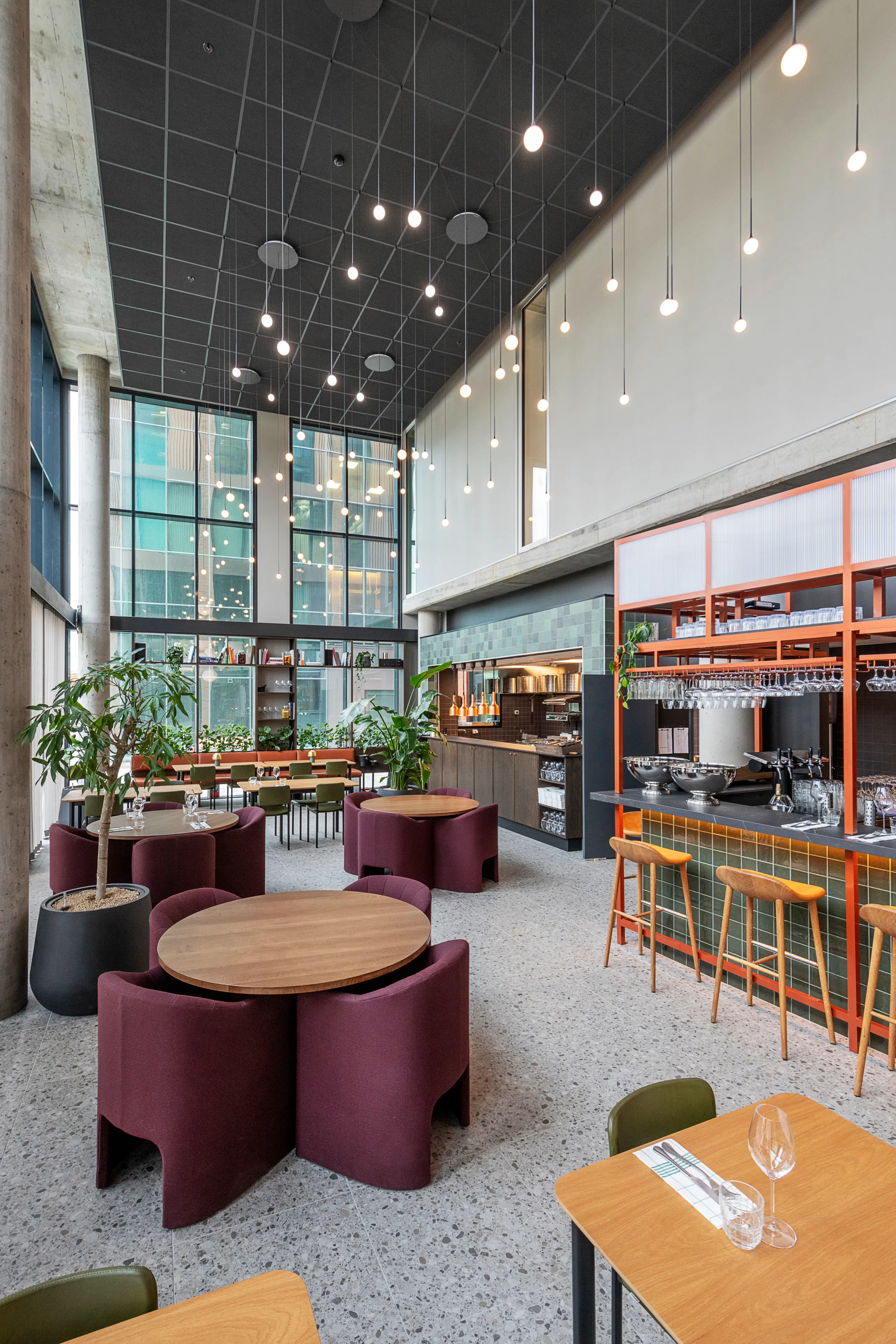 Modern office breakout area interior with high ceilings, black acoustic panels, hanging pendant lights, round tables with burgundy seating, and a bar area with wooden stools and green tiles