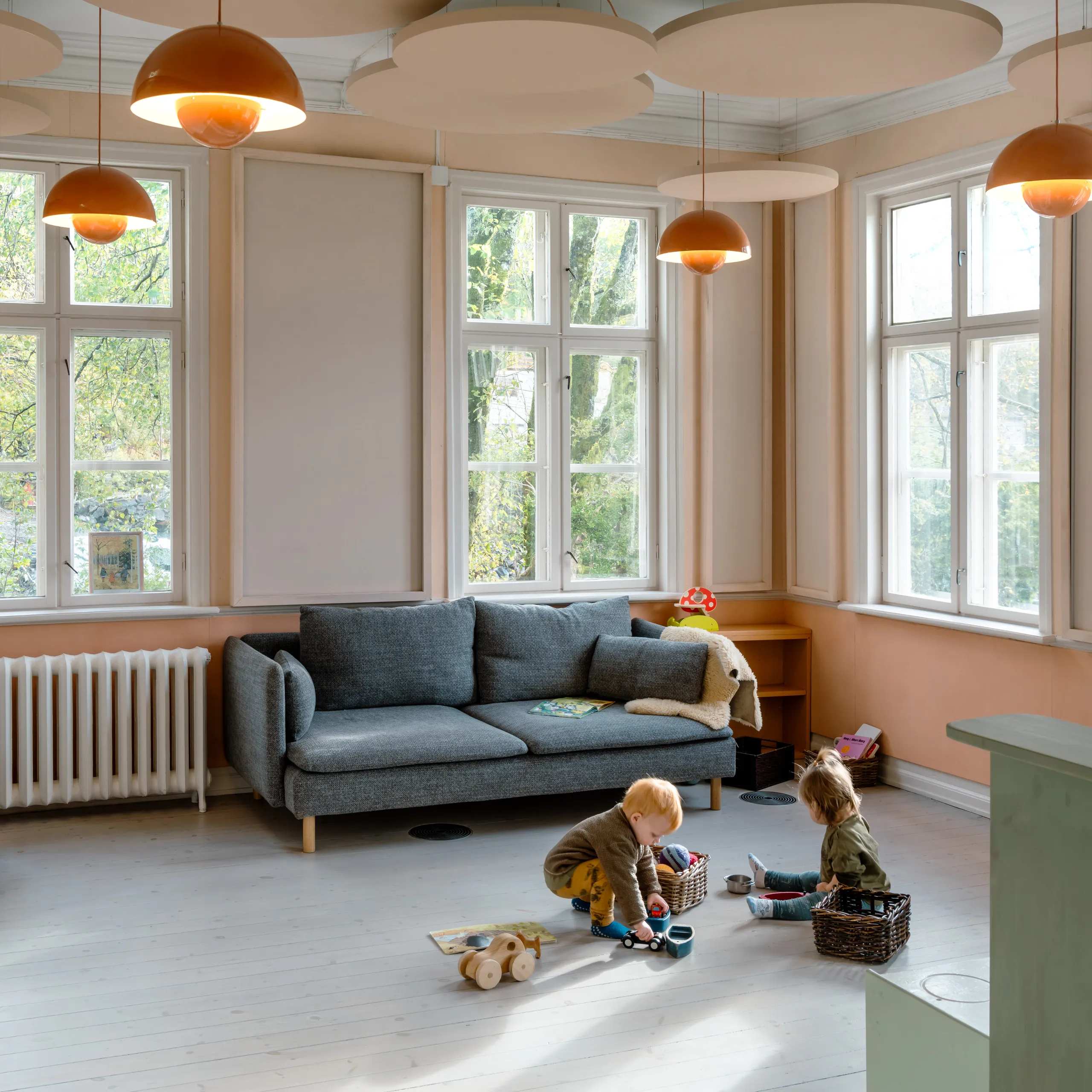 A bright children's playroom featuring suspended circular acoustic ceiling islands and orange pendant lights, with large windows, a grey sofa, and two toddlers playing on the wooden floor.