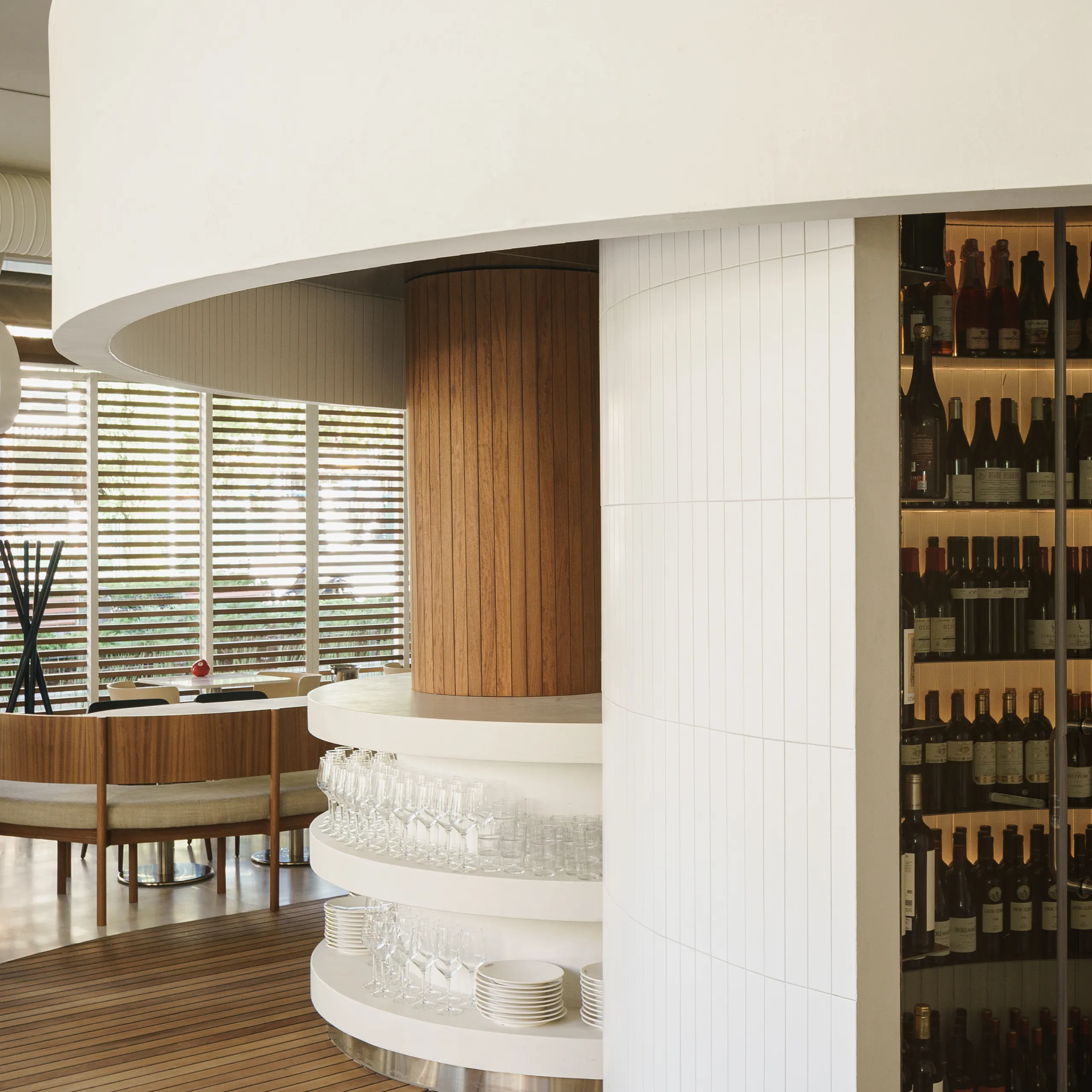Restaurant interior with a curved monolithic acoustic ceiling above a circular bar, featuring wood columns, wine display shelving, and seating near large windows.
