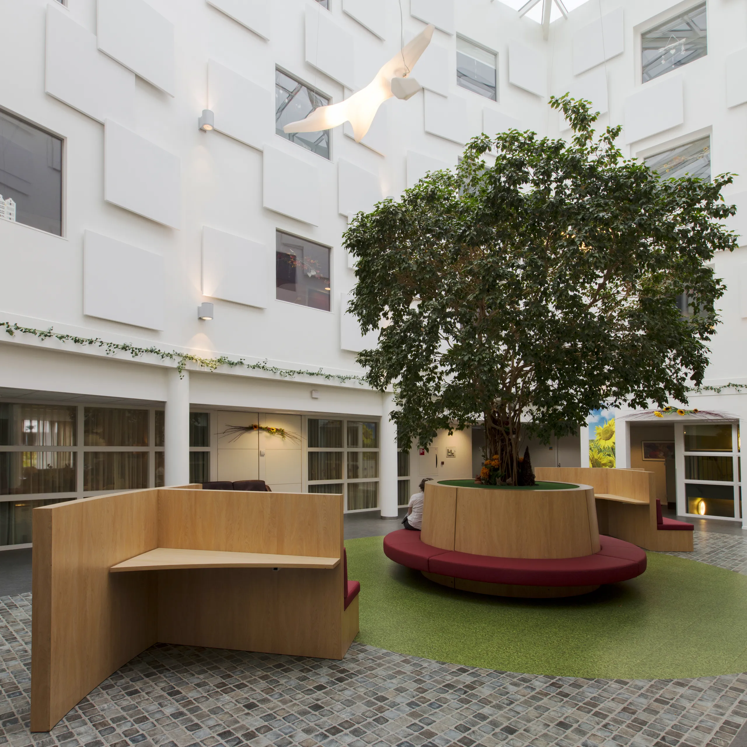 A bright multi-storey atrium featuring staggered white acoustic wall panels and a skylight roof, with seating positioned around a central tree.