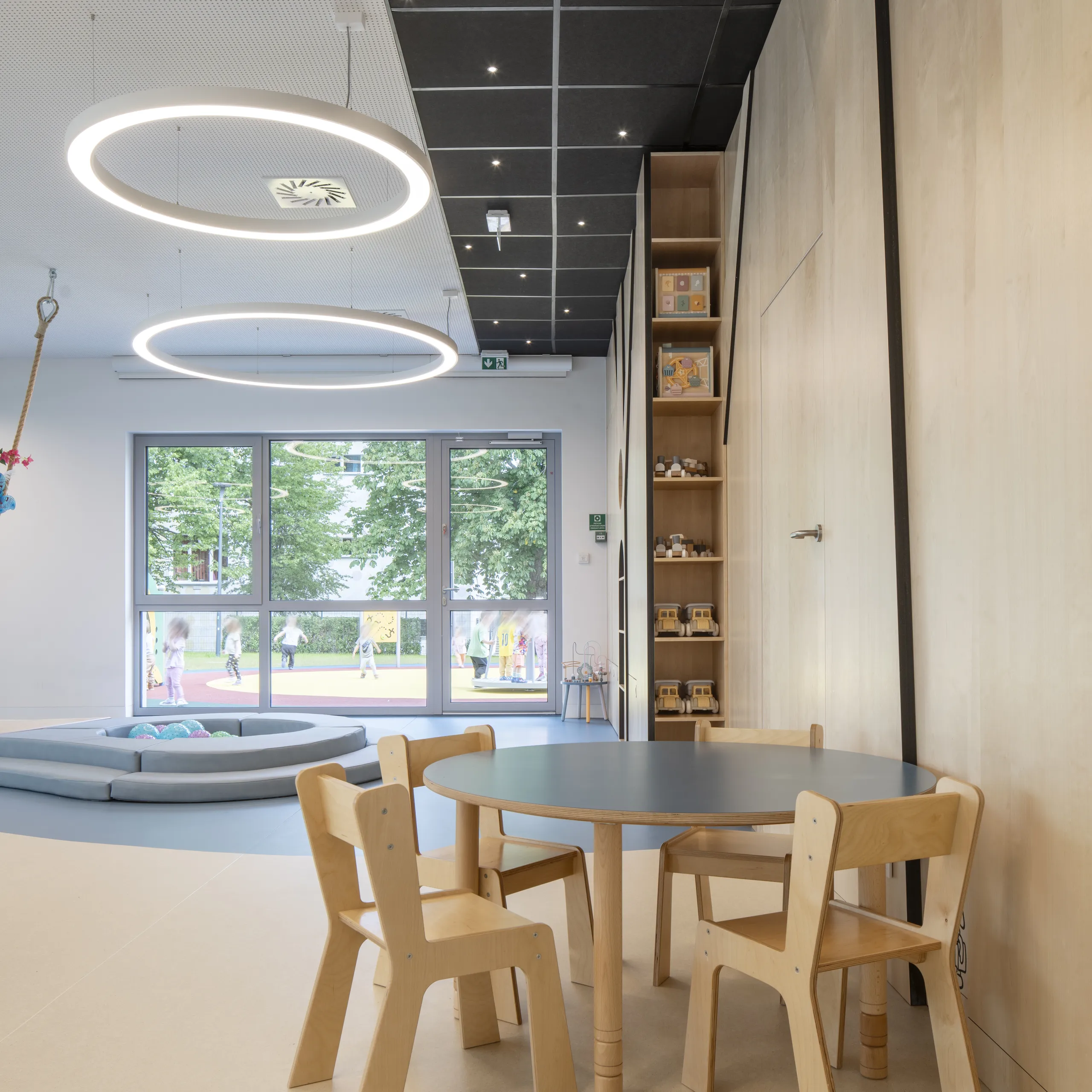 A light-filled playroom featuring black acoustic ceiling tiles with tiny integrated spotlights, large circular pendant lights, and a children’s table set beside tall windows.