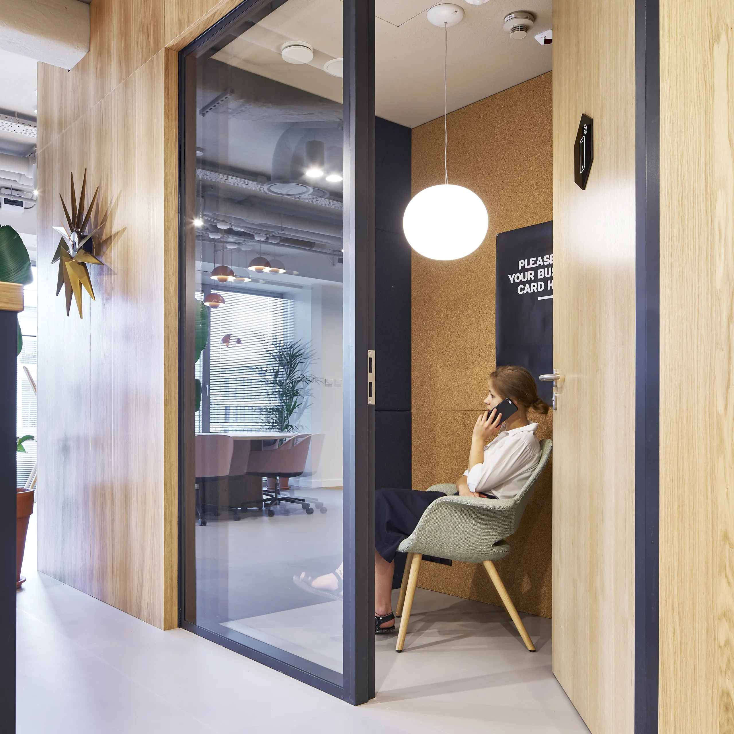 Phone booth area with acoustic ceiling panels and wooden walls, where a person sits in a lounge chair making a call