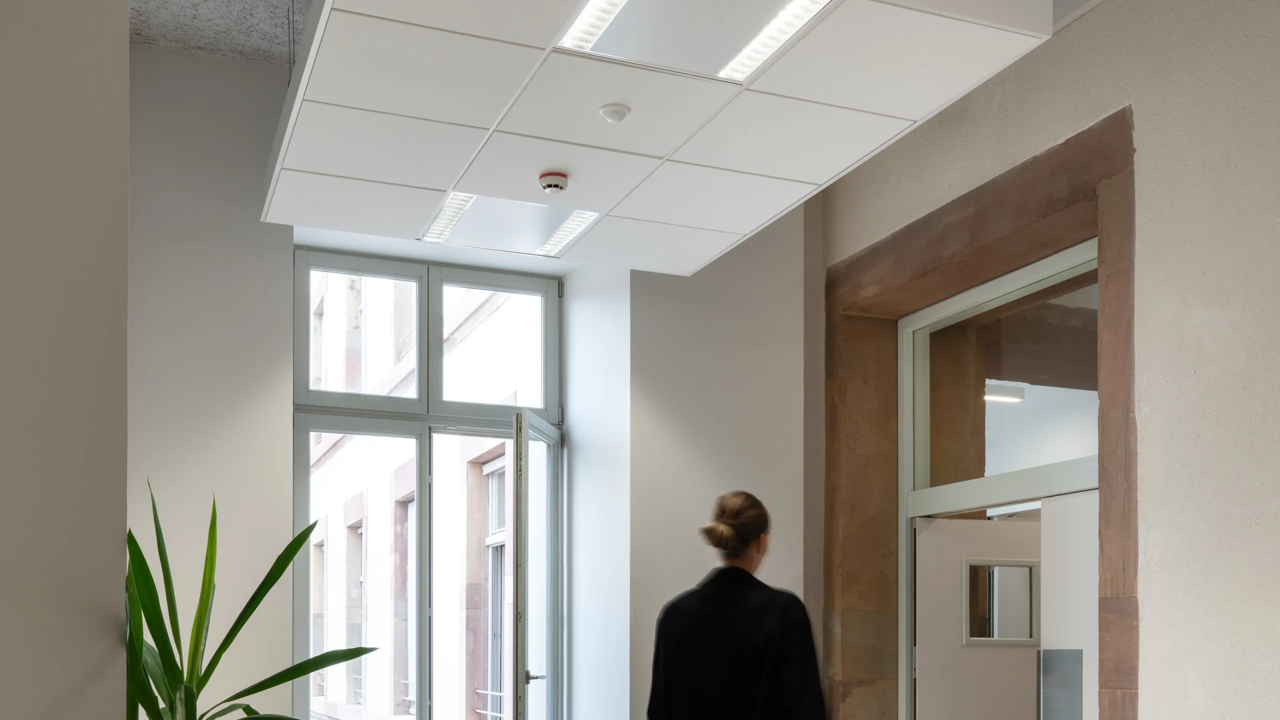 Person in black outfit walking in a modern, minimalist office hallway with a large window and floating raft ceiling