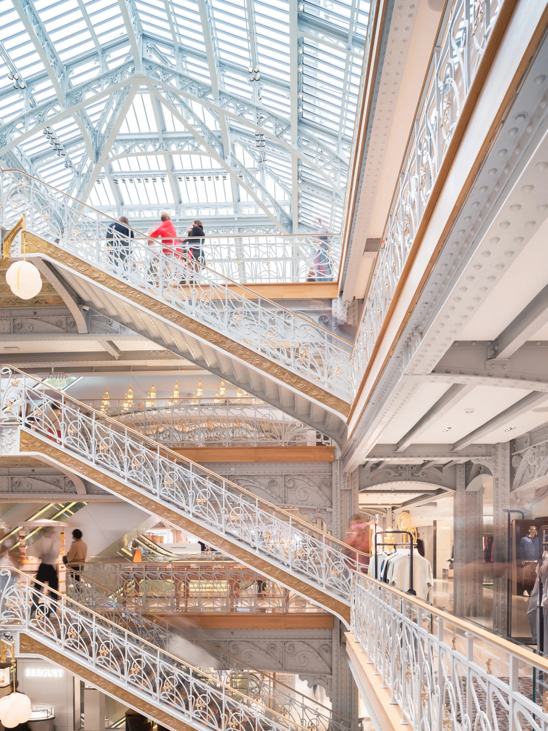 Interior of a multi-level department store with ornate staircases, decorative metal railings, and a large glass roof bringing daylight into the atrium.