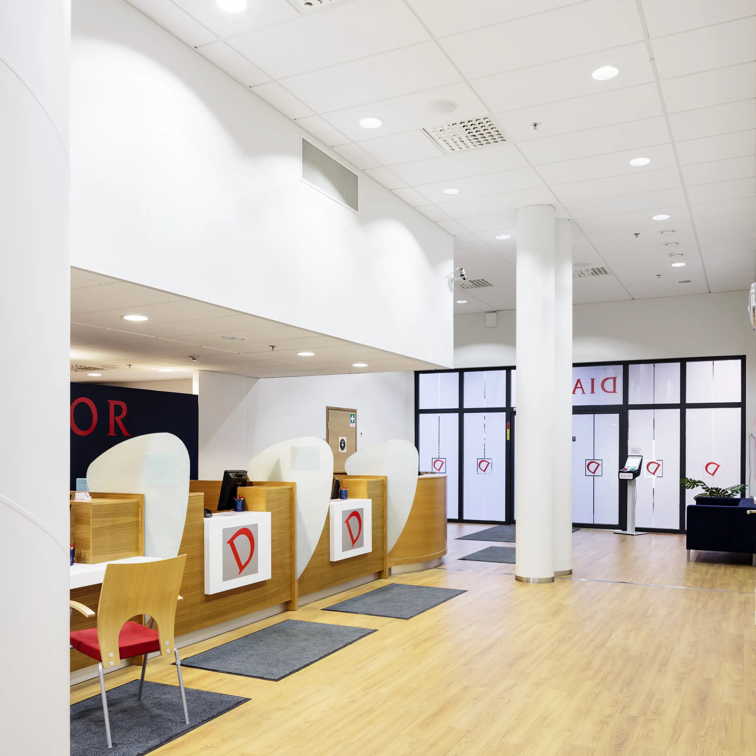 A bright healthcare reception area with wood-panelled service counters, red signage, white acoustic ceiling tiles and recessed downlights.
