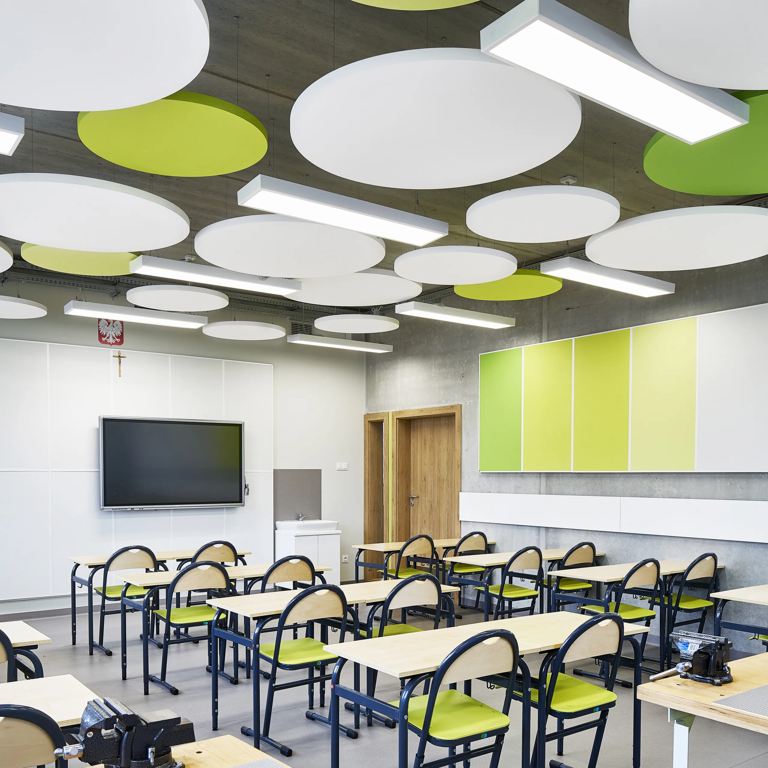 A bright classroom featuring suspended circular acoustic islands in white and lime green, complementing the exposed concrete ceiling and coordinated wall panels.