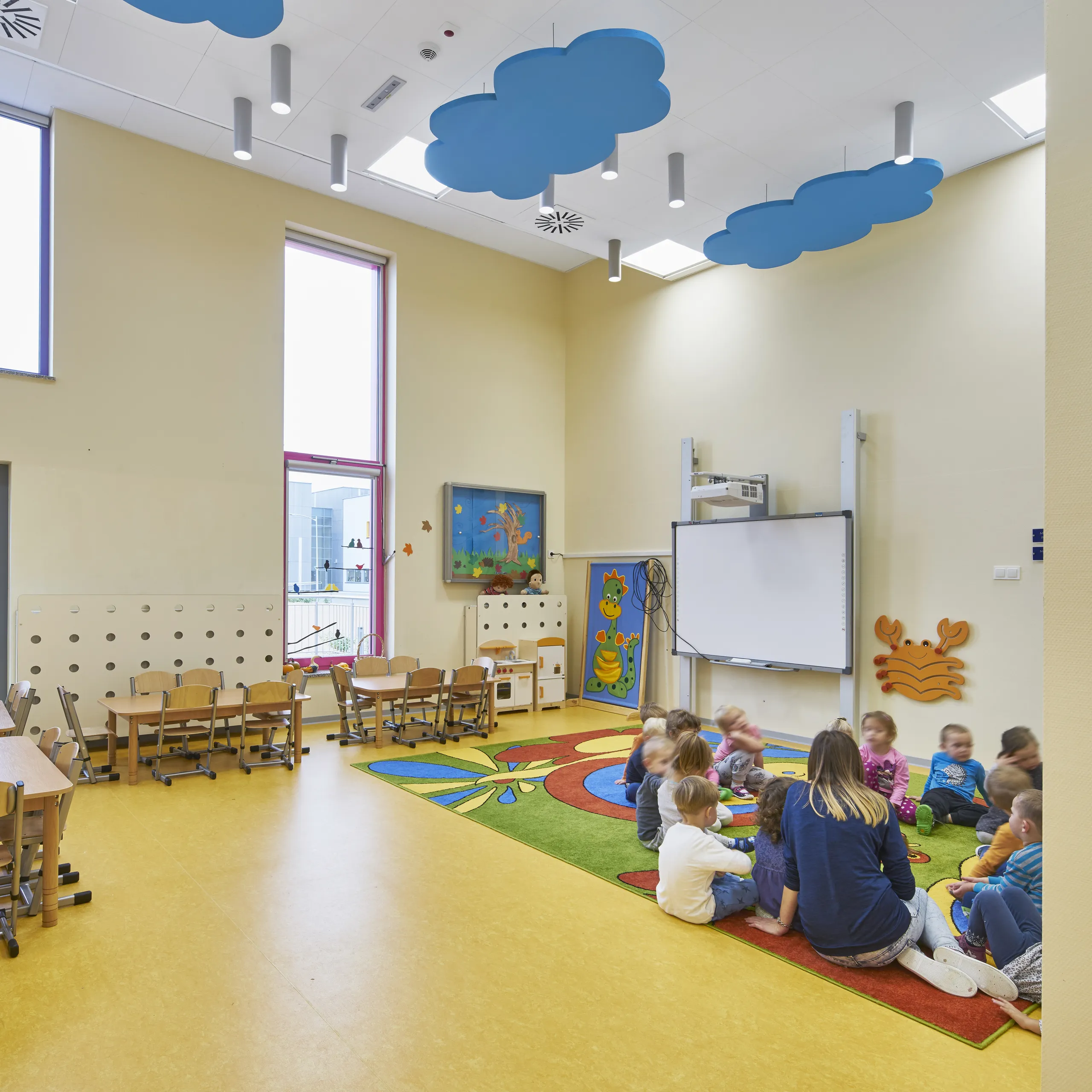 A playful nursery classroom featuring blue cloud-shaped acoustic ceiling islands suspended under white acoustic ceiling tiles and cylindrical pendant lights, with young children gathered on a colourful rug beneath high windows.