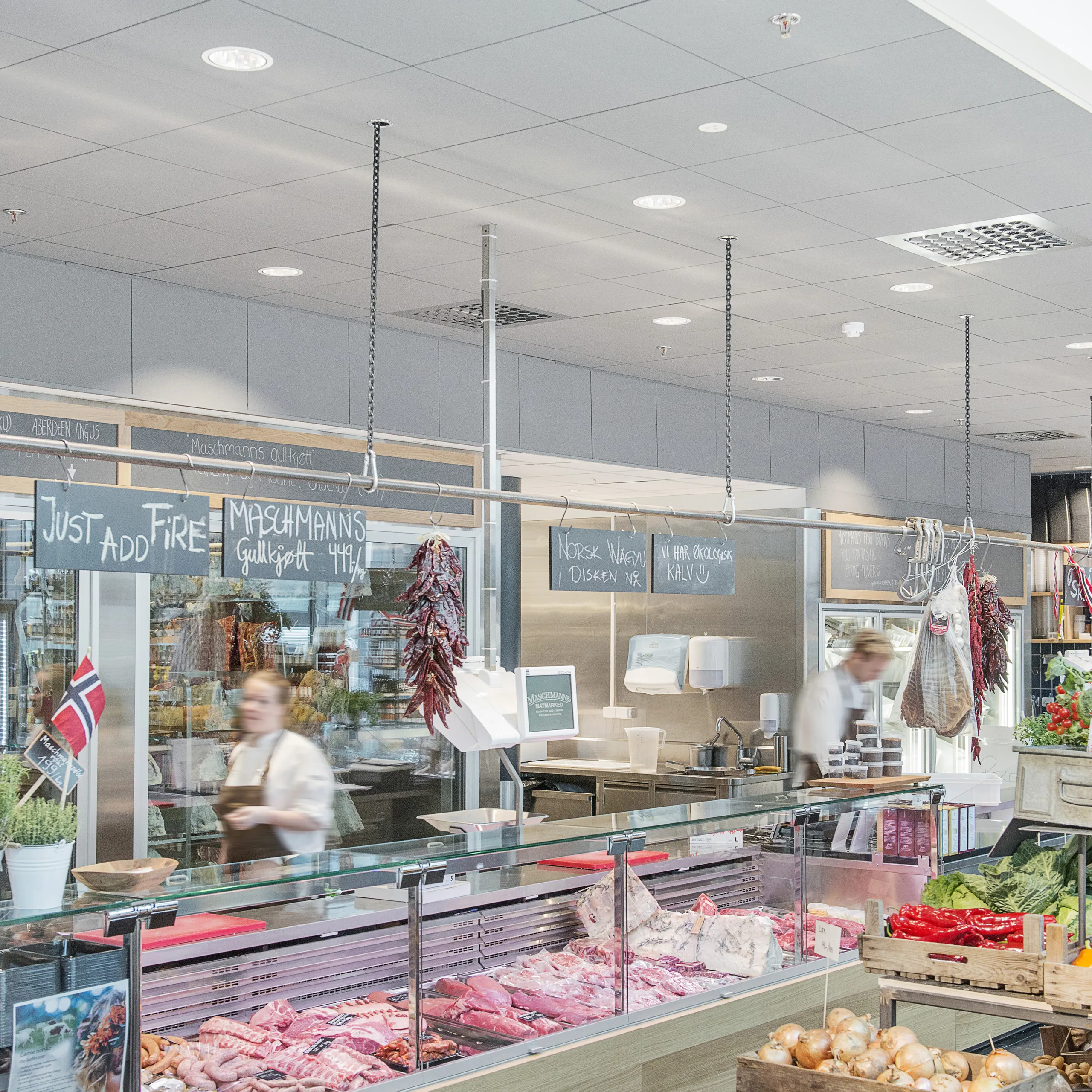 Butcher counter at a food market with refrigerated meat displays and a grey acoustic tile ceiling above the service area.