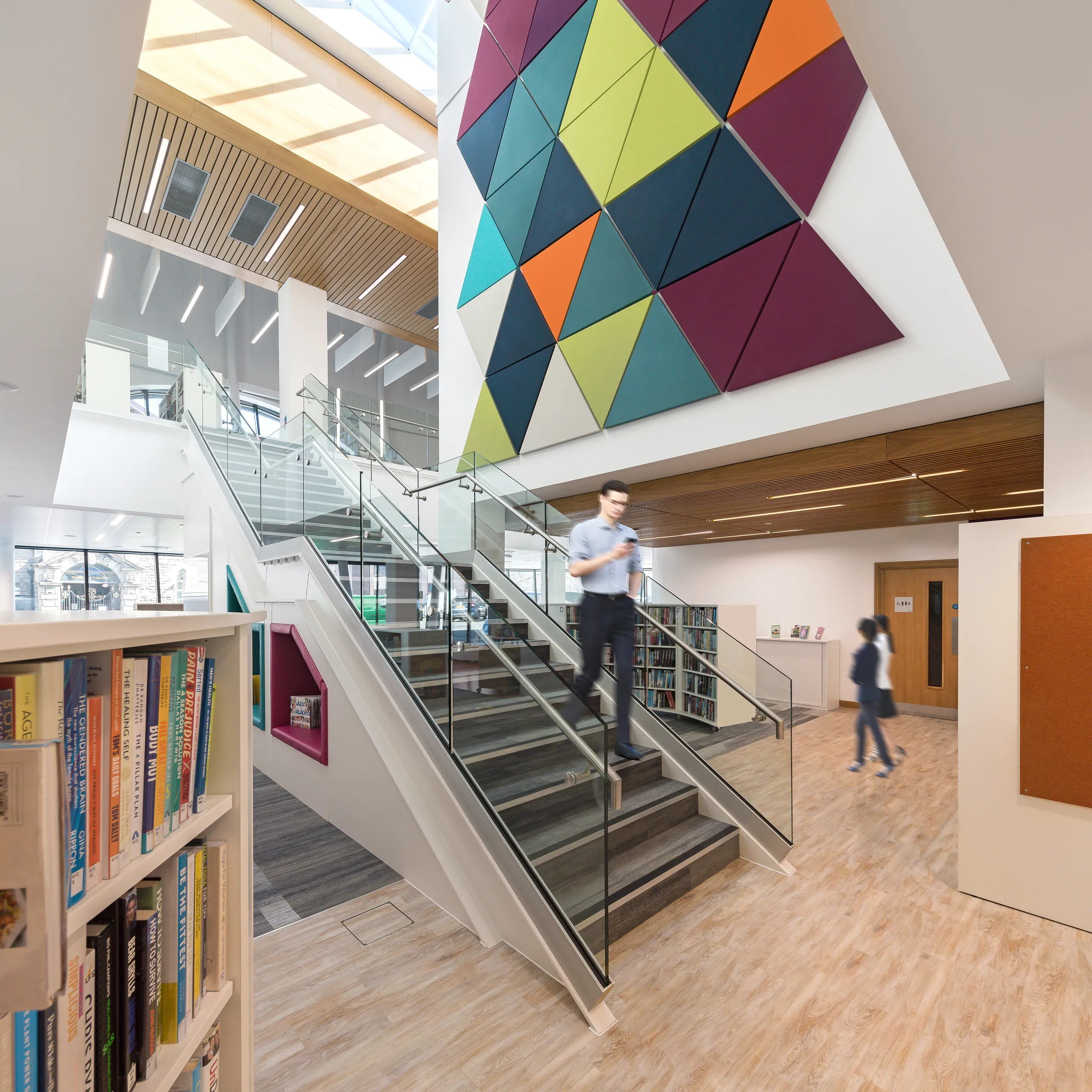 Library atrium with a staircase, bookshelves, and a large colourful geometric acoustic wall panel installation beneath a skylight.