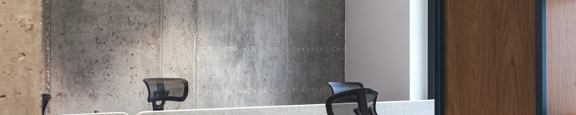 Open-plan office with suspended acoustic ceiling panels, exposed ductwork painted blue, concrete wall finish, and modern workstations with grey dividers