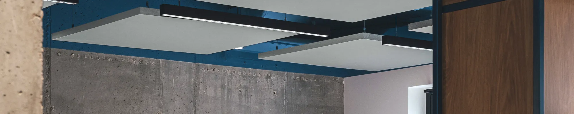 Open-plan office with suspended acoustic ceiling panels, exposed ductwork painted blue, concrete wall finish, and modern workstations with grey dividers