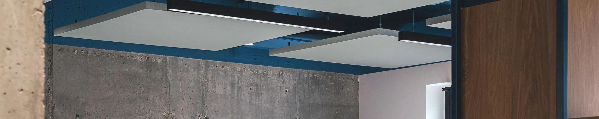 Open-plan office with suspended acoustic ceiling panels, exposed ductwork painted blue, concrete wall finish, and modern workstations with grey dividers