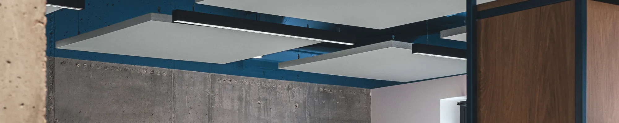 Open-plan office with suspended acoustic ceiling panels, exposed ductwork painted blue, concrete wall finish, and modern workstations with grey dividers