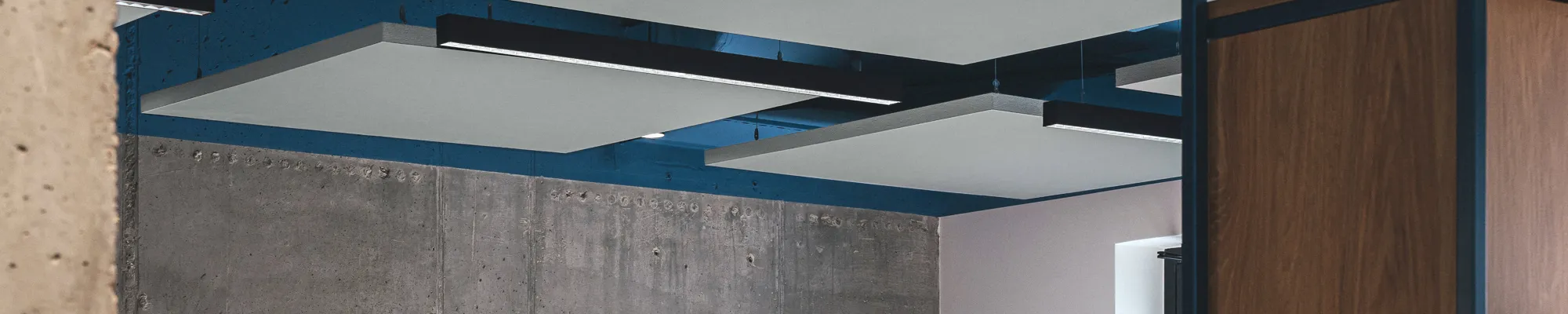 Open-plan office with suspended acoustic ceiling panels, exposed ductwork painted blue, concrete wall finish, and modern workstations with grey dividers