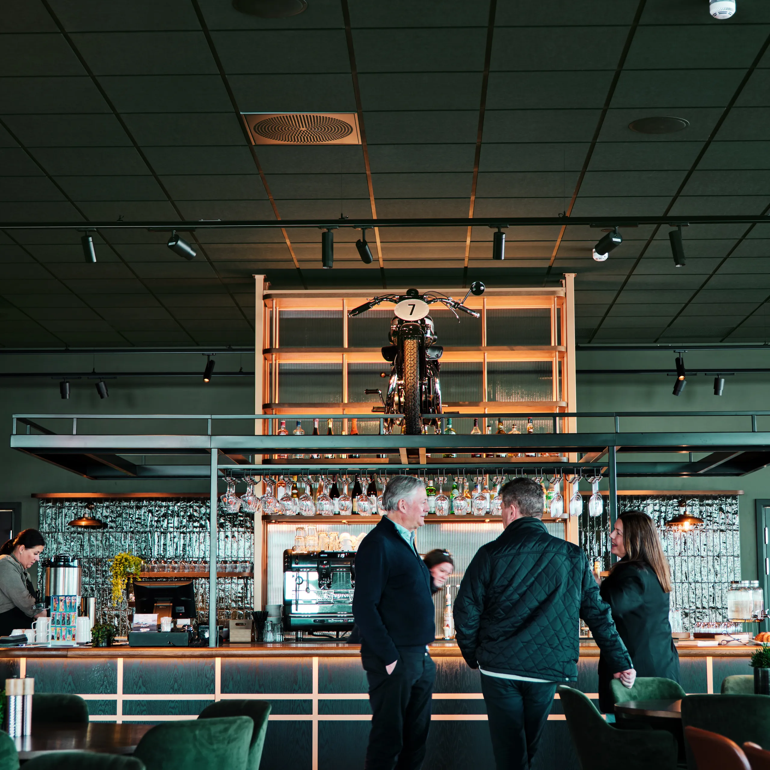 Restaurant interior with a dark acoustic ceiling grid and track lighting above a bar counter, where three people stand talking beneath a display shelf holding a motorcycle.