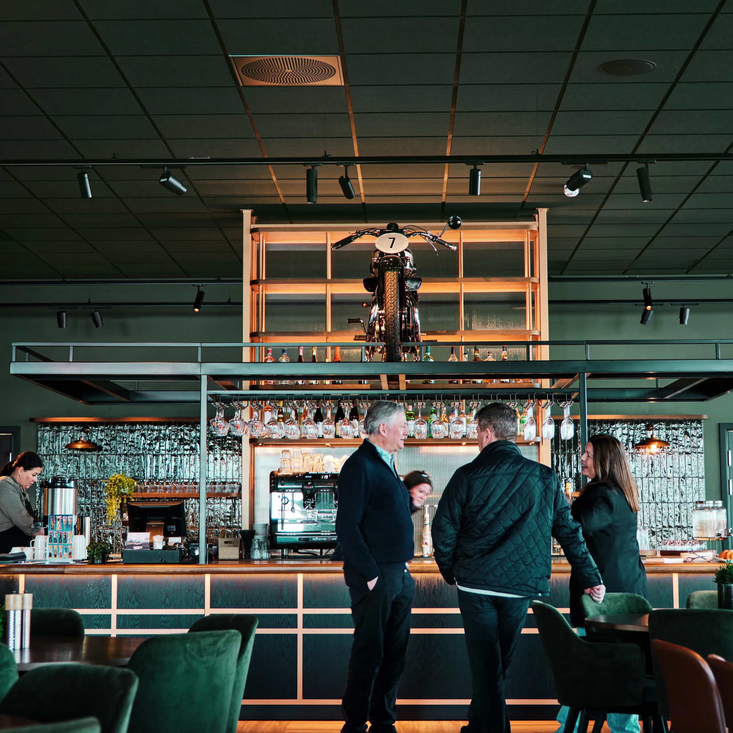 A modern bar interior featuring dark green acoustic ceiling tiles, track lighting, and a display shelf holding a vintage motorcycle above the bar counter. People are gathered at the bar, surrounded by dark green seating and warm wooden finishes.