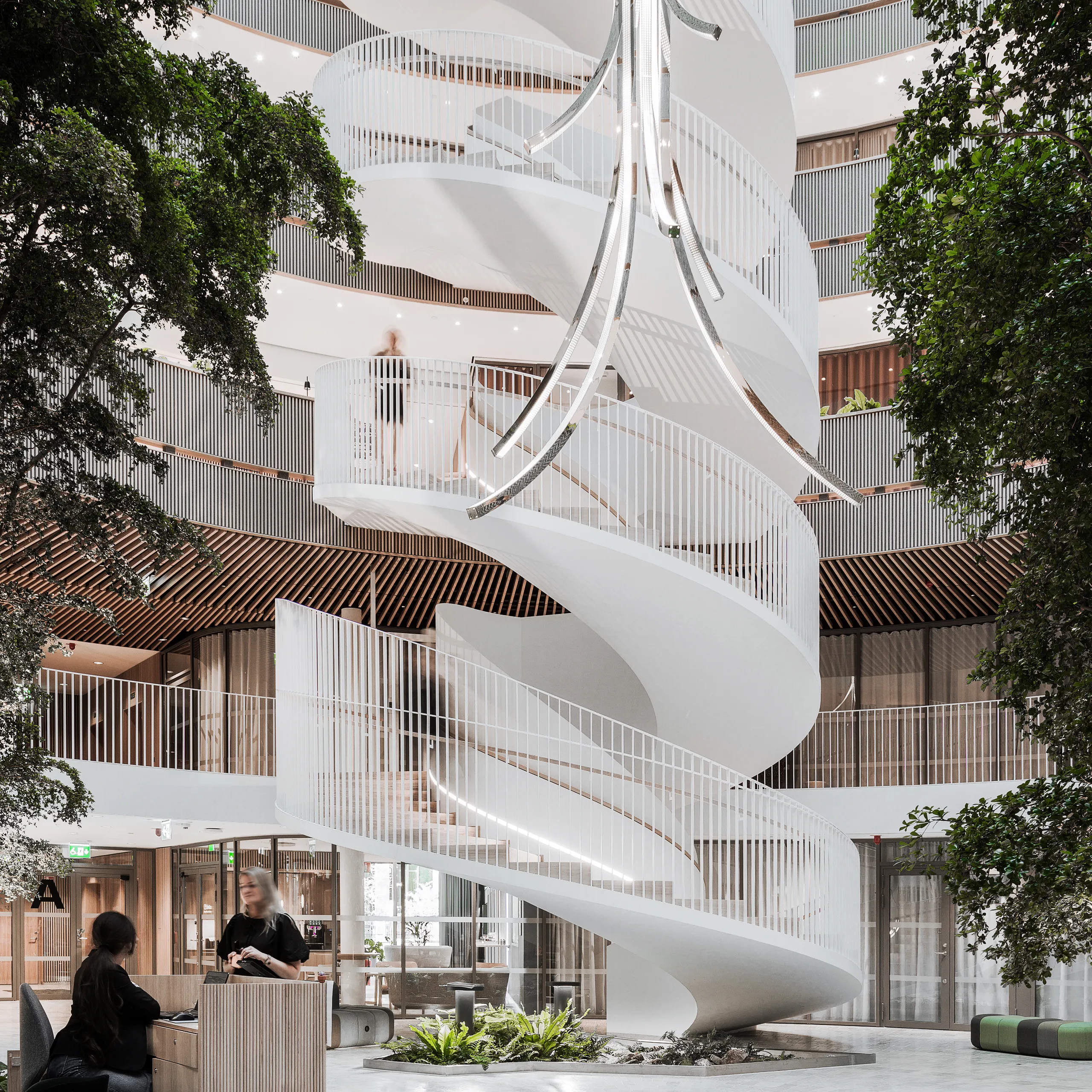 Reception area in a multi-storey atrium with a monolithic acoustic ceiling and monolithic acoustic walls, featuring a sculptural white spiral staircase and large indoor trees