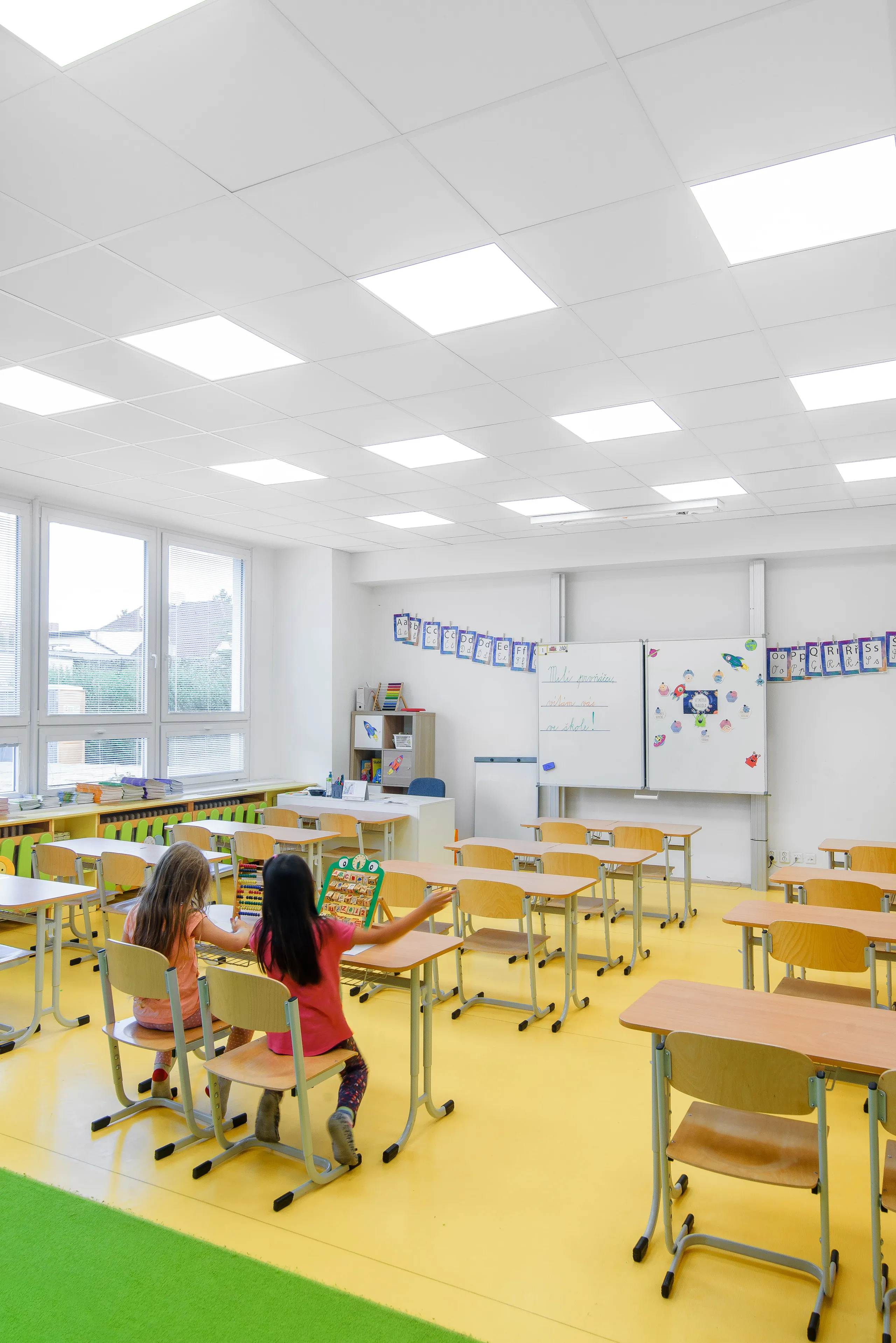 A bright and tidy primary classroom featuring a white acoustic ceiling grid, large windows, colourful teaching displays, and two children working together at the front of the room