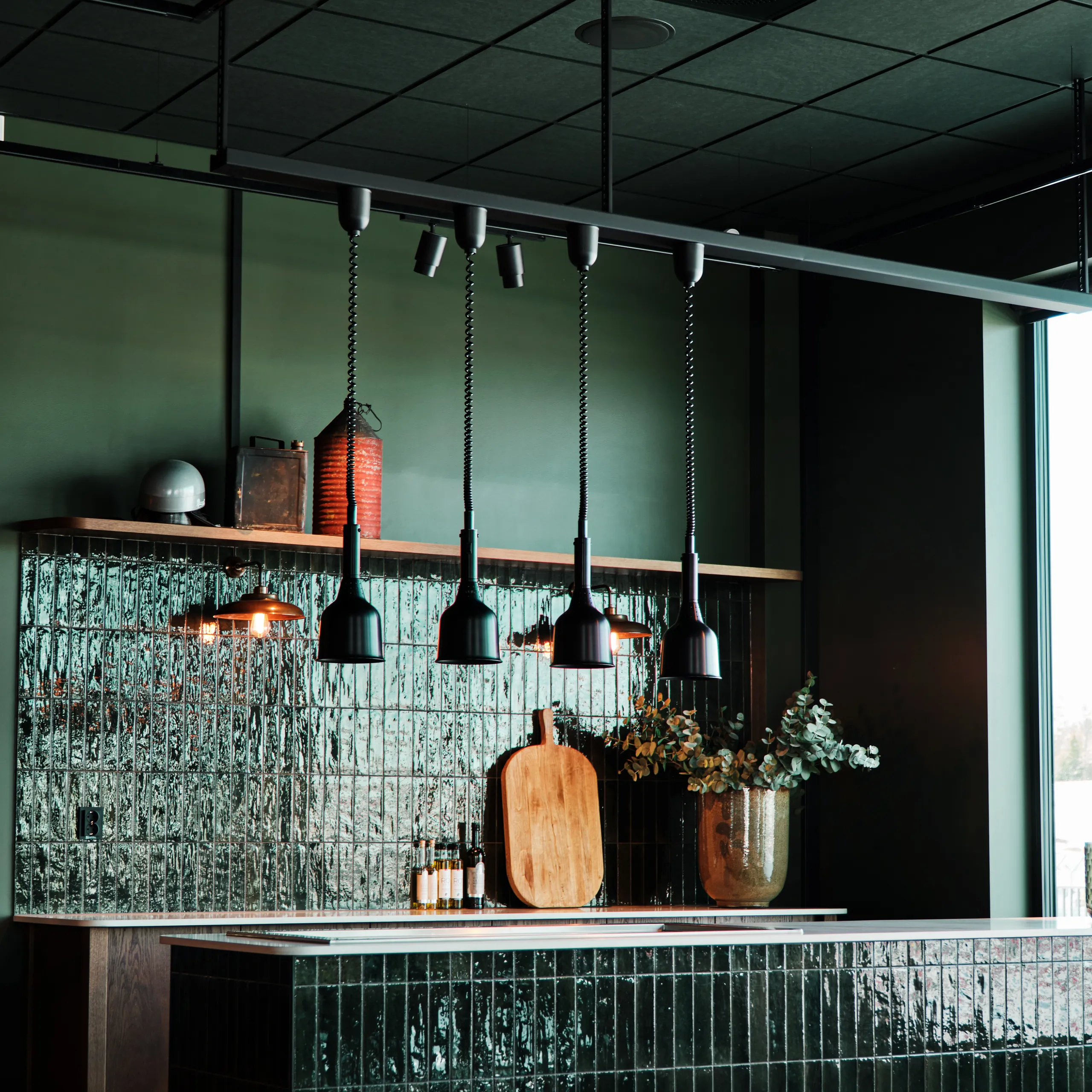 Modern kitchen with dark green tiled island, pendant lights, wooden cutting boards, and a vase with plants, set against textured walls.