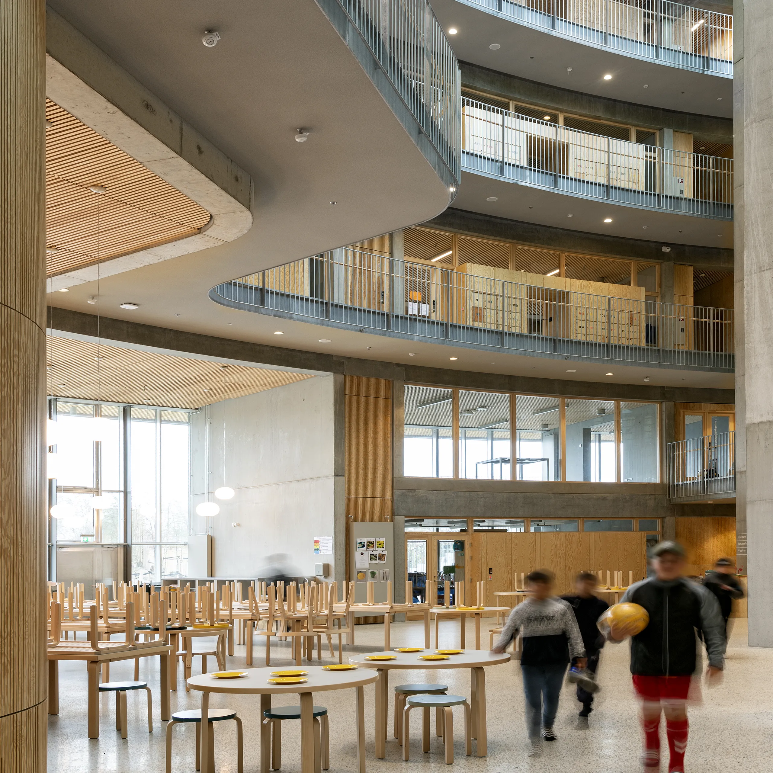 A spacious multi-storey school atrium featuring a seamless monolithic grey acoustic ceiling, curved balcony edges, timber details, and students moving through the bright central space.