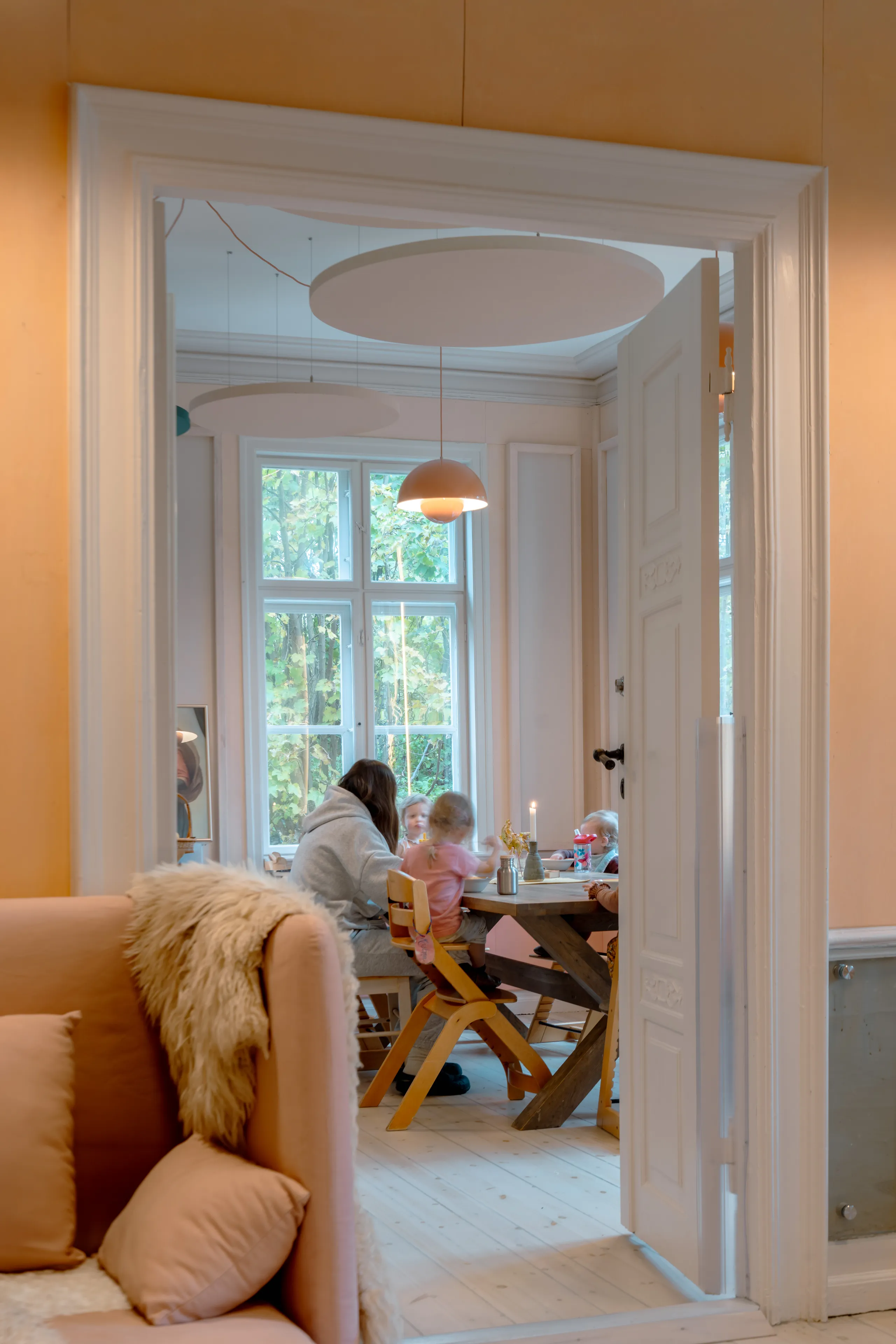 A soft, light-filled playroom where circular acoustic ceiling islands and warm pendant lights create a calm setting as children play beneath large windows
