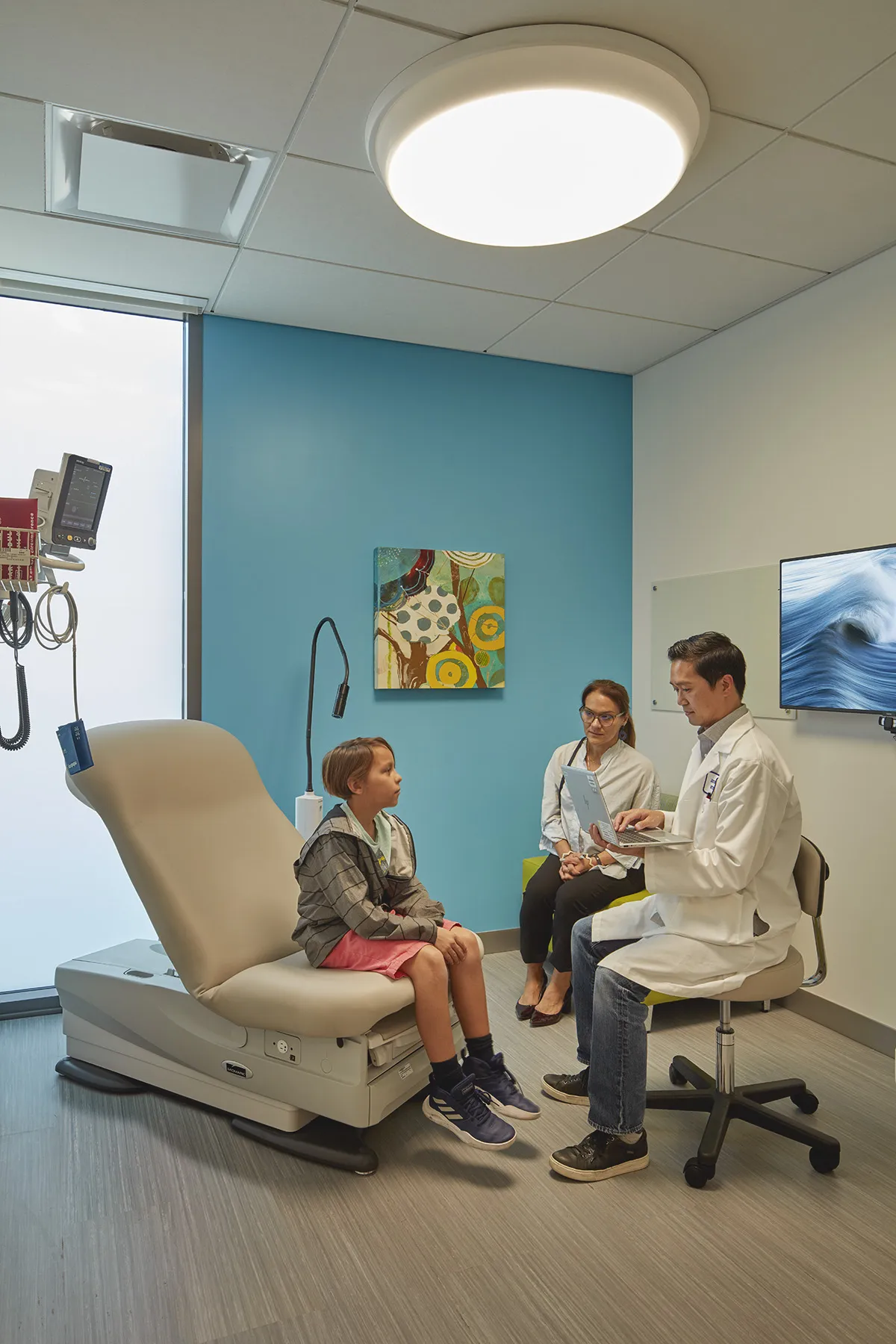 A paediatric examination room with a doctor speaking to a child and guardian, showing a white acoustic ceiling with integrated lighting and medical equipment.