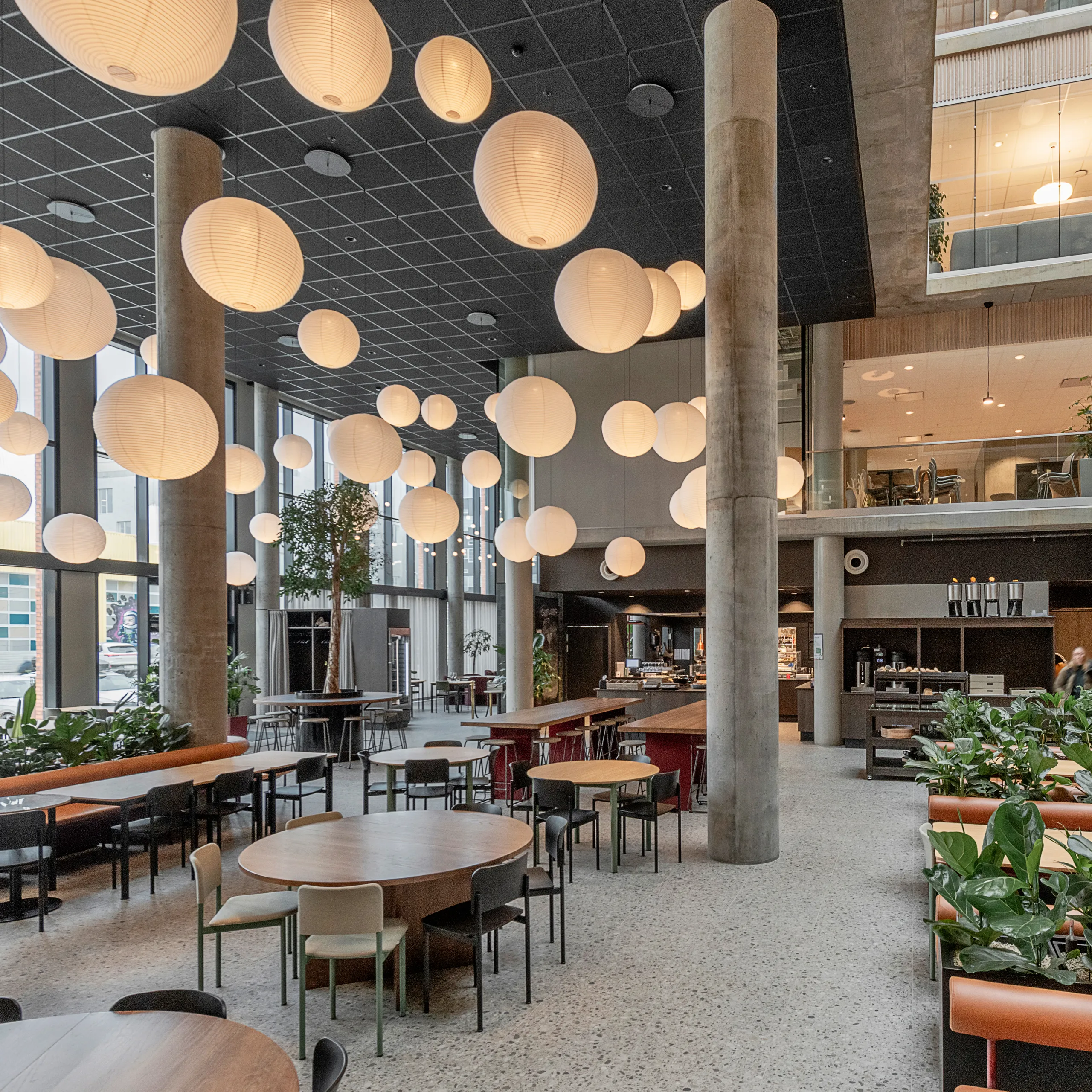 Atrium dining area with dark acoustic ceiling tiles, suspended globe pendant lights, tall columns, and seating areas arranged around planting and open social spaces.