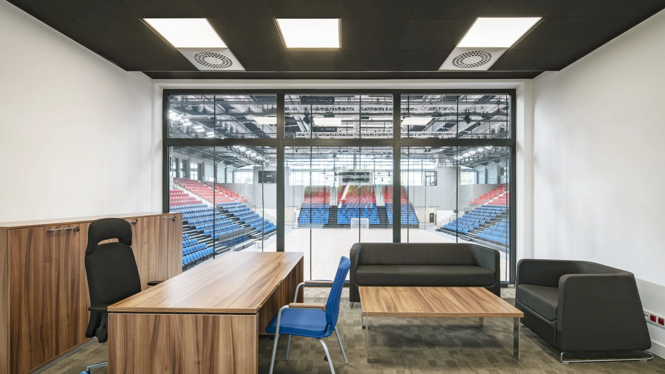 Office with black suspended ceiling, wooden desk, chair, and black sofas overlooks an indoor sports arena with red and blue seating.