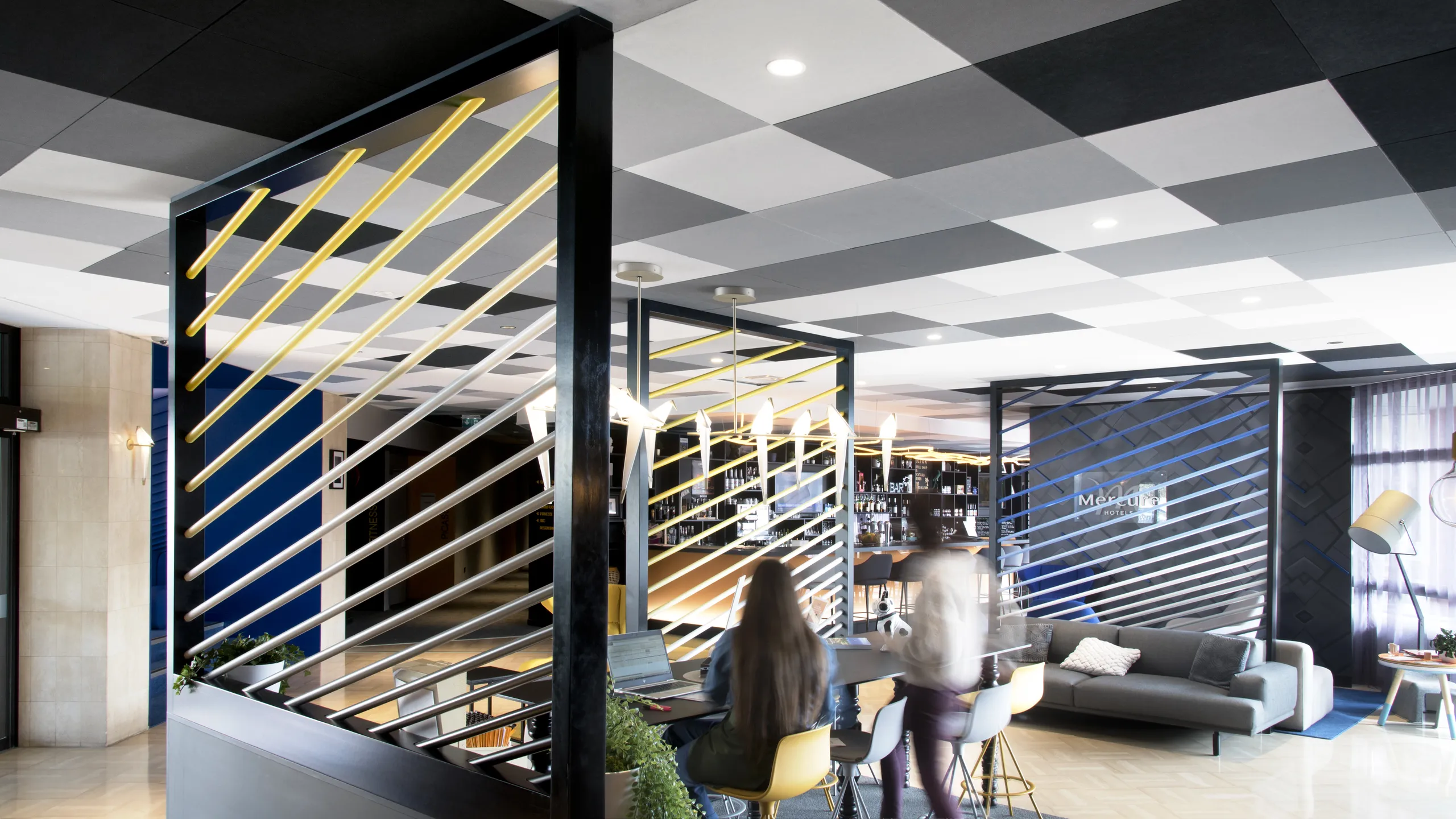 Hotel bar and lounge with a black and white checkerboard acoustic ceiling, illuminated bar counter, seating areas, and large windows bringing daylight into the space.