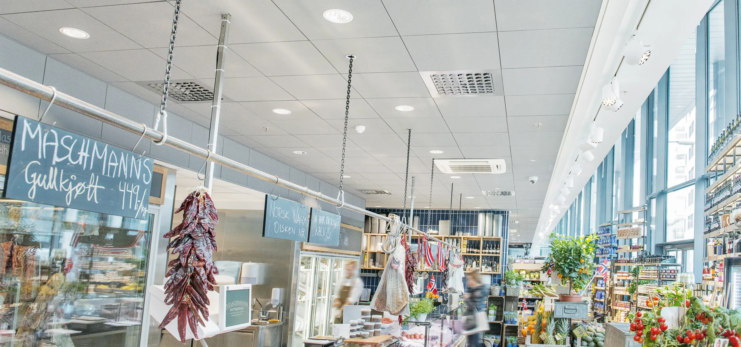 Food court interior with grey acoustic ceiling tiles, pendant lighting, open food counter, and dining area with tables and seated guests in a bright, busy setting.