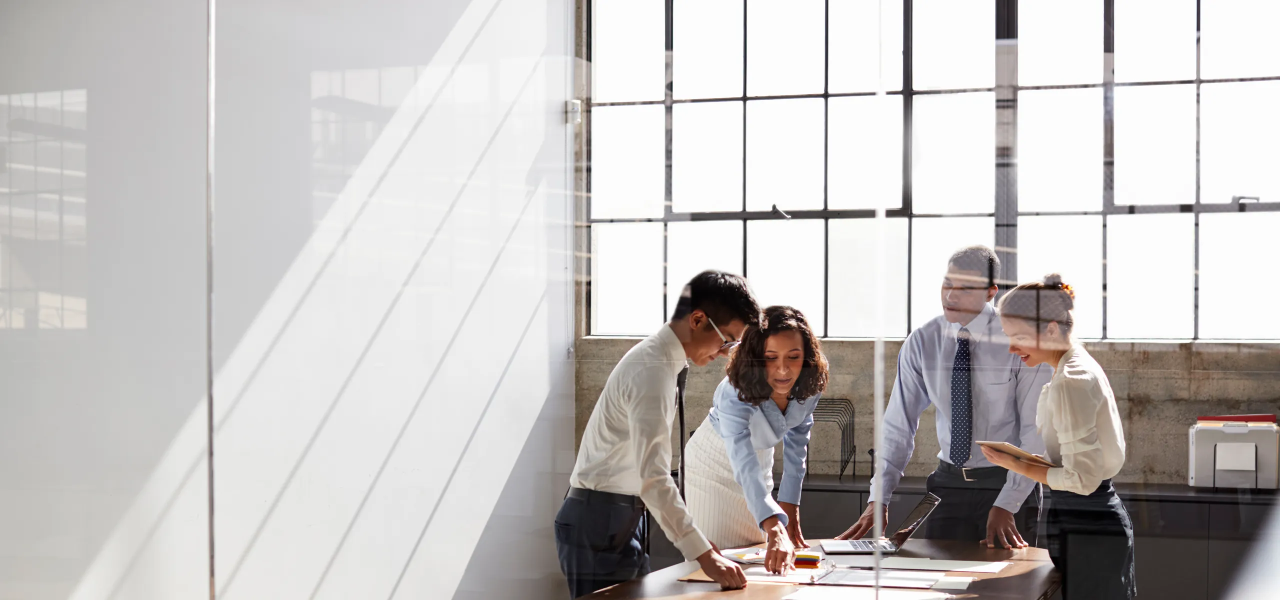 Modern office meeting with four professionals working together at a table in a loft-style room with tall steel-framed windows