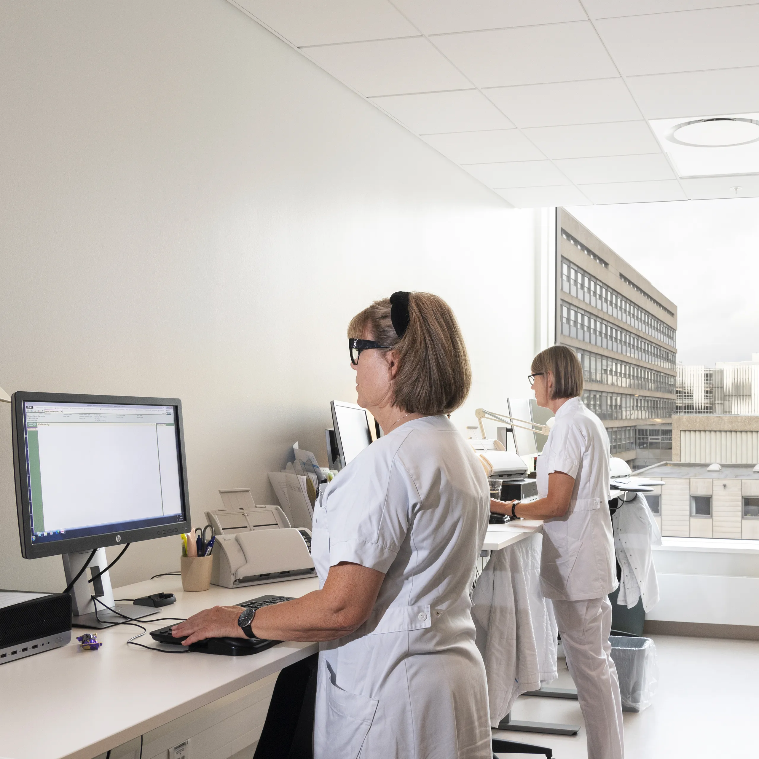 Two healthcare staff working at computers in a bright clinical office with a white acoustic ceiling and large window overlooking adjacent hospital buildings.