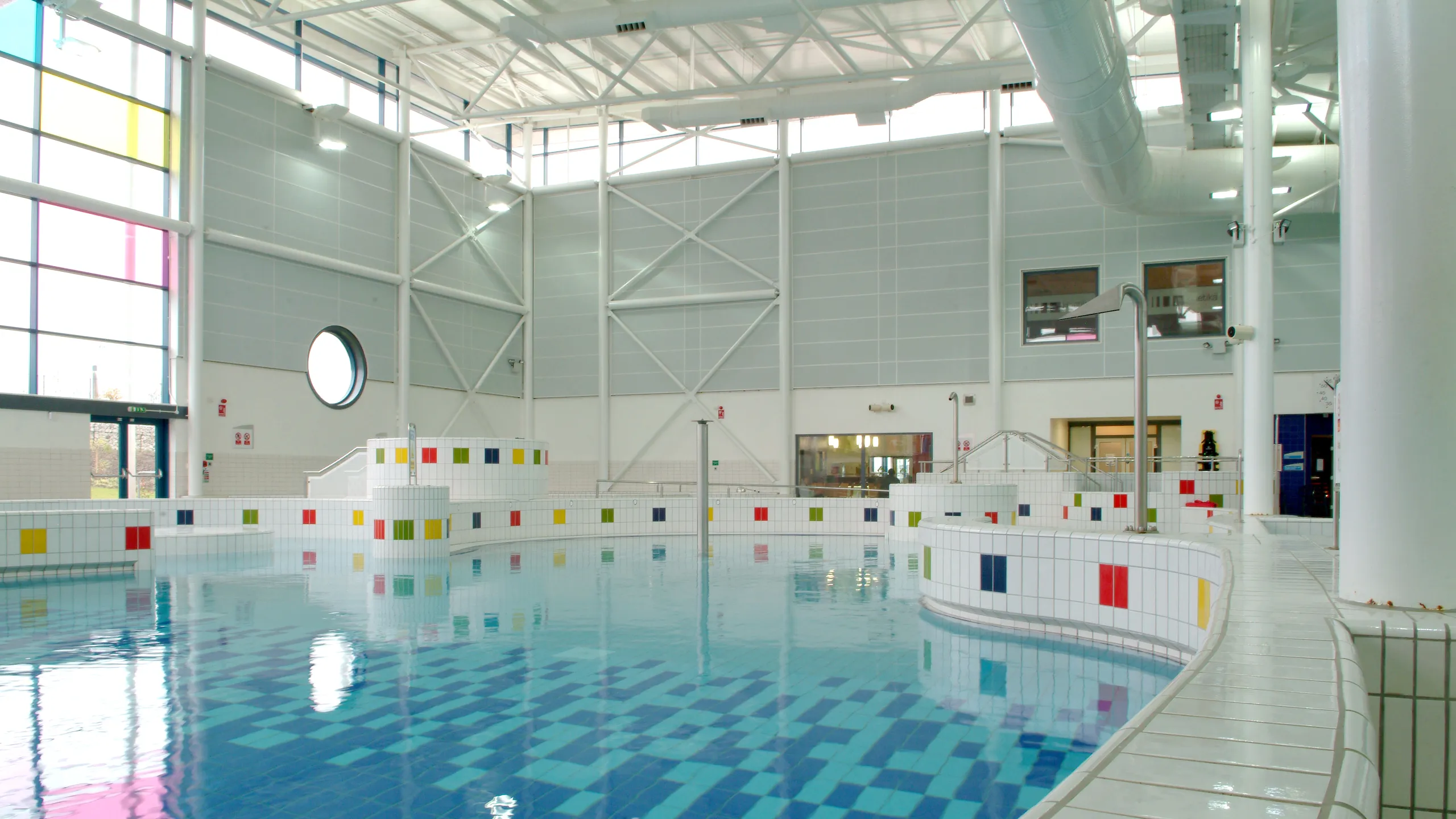 Indoor leisure pool with Rockfon Color-all wall absorbers, tiled pool edges with coloured accents, and a bright, open hall with large windows and high ceilings.
