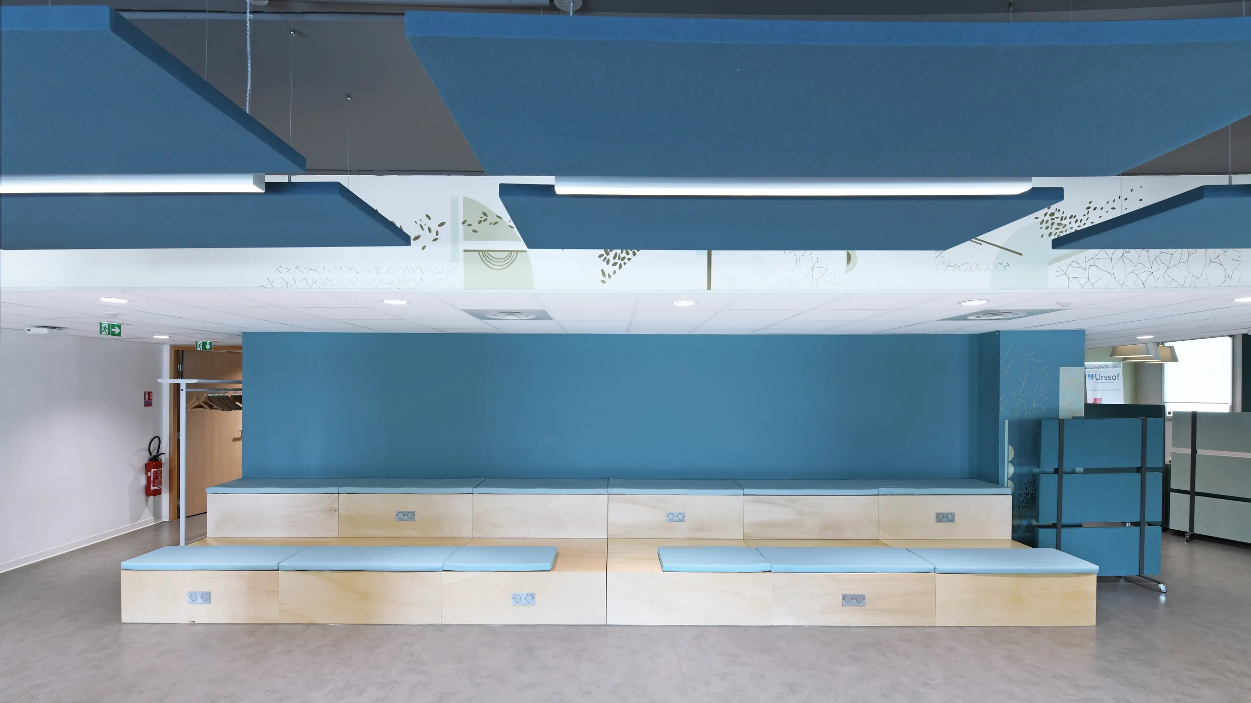 Modern waiting area with blue floating ceiling panels, light wood benches, blue accent wall, and decorative ceiling panels. Door and emergency exit signs are visible.