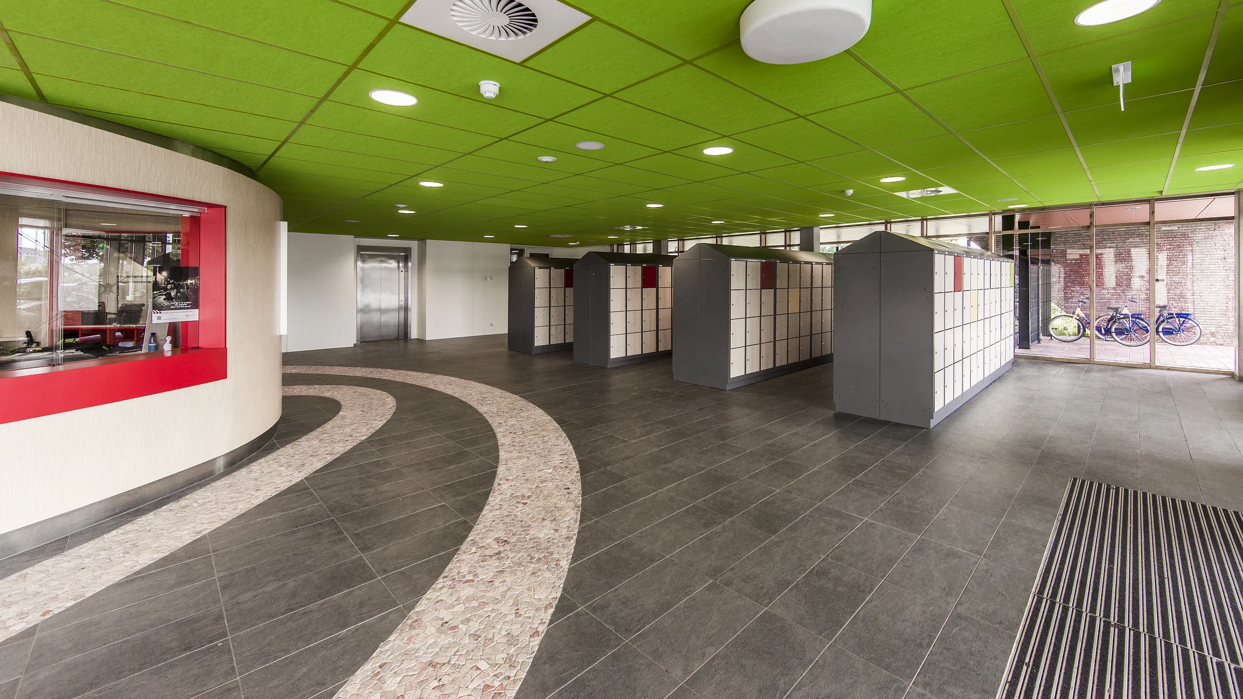 Modern lobby with a green ceiling, red accents, and a curved pathway. Glass doors, elevators, and bike racks are visible in the background.
