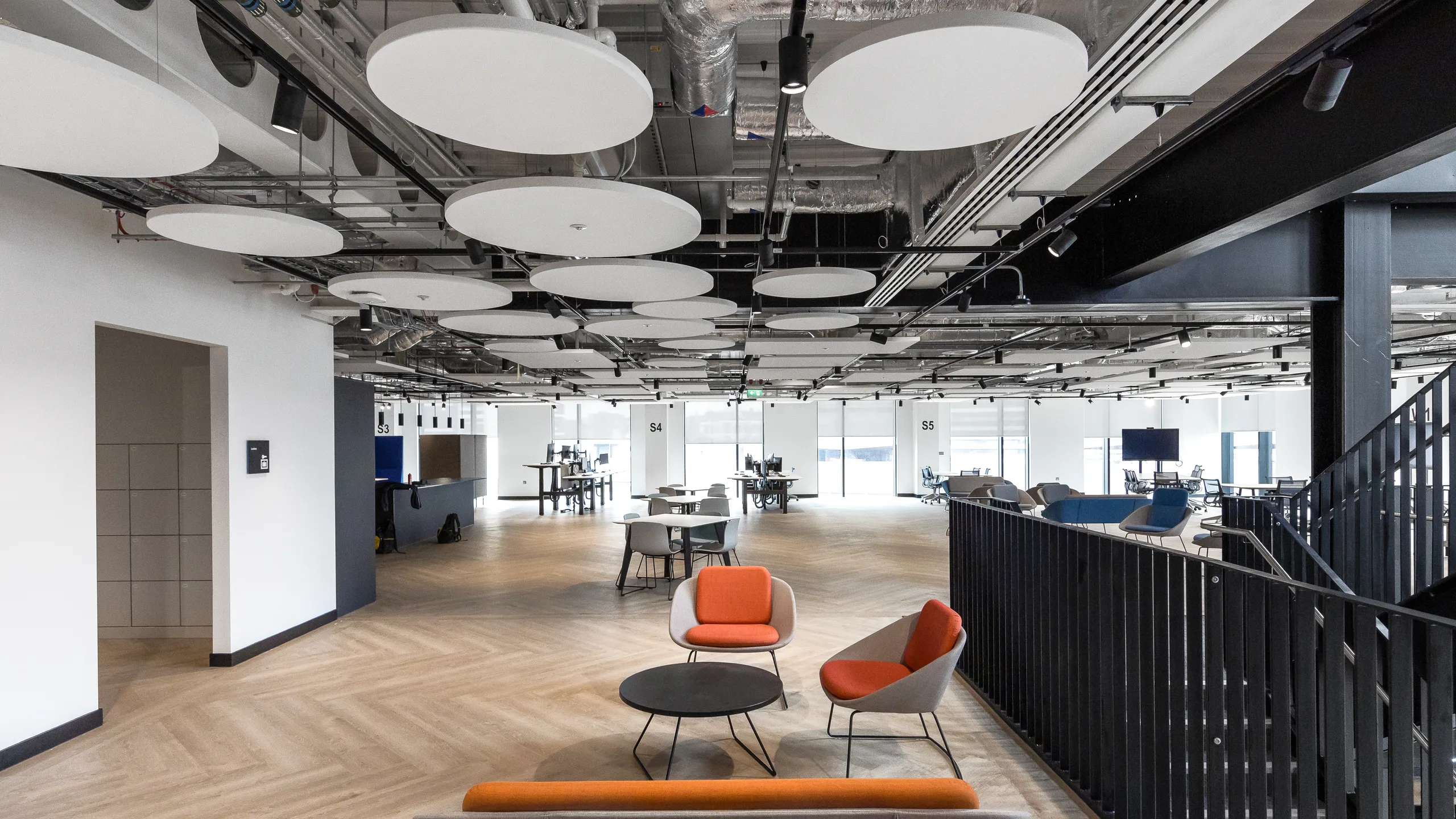 Modern office space with an office ceiling composed of floating circular ceiling panels, open seating area featuring orange and gray chairs, large windows, and wooden flooring.
