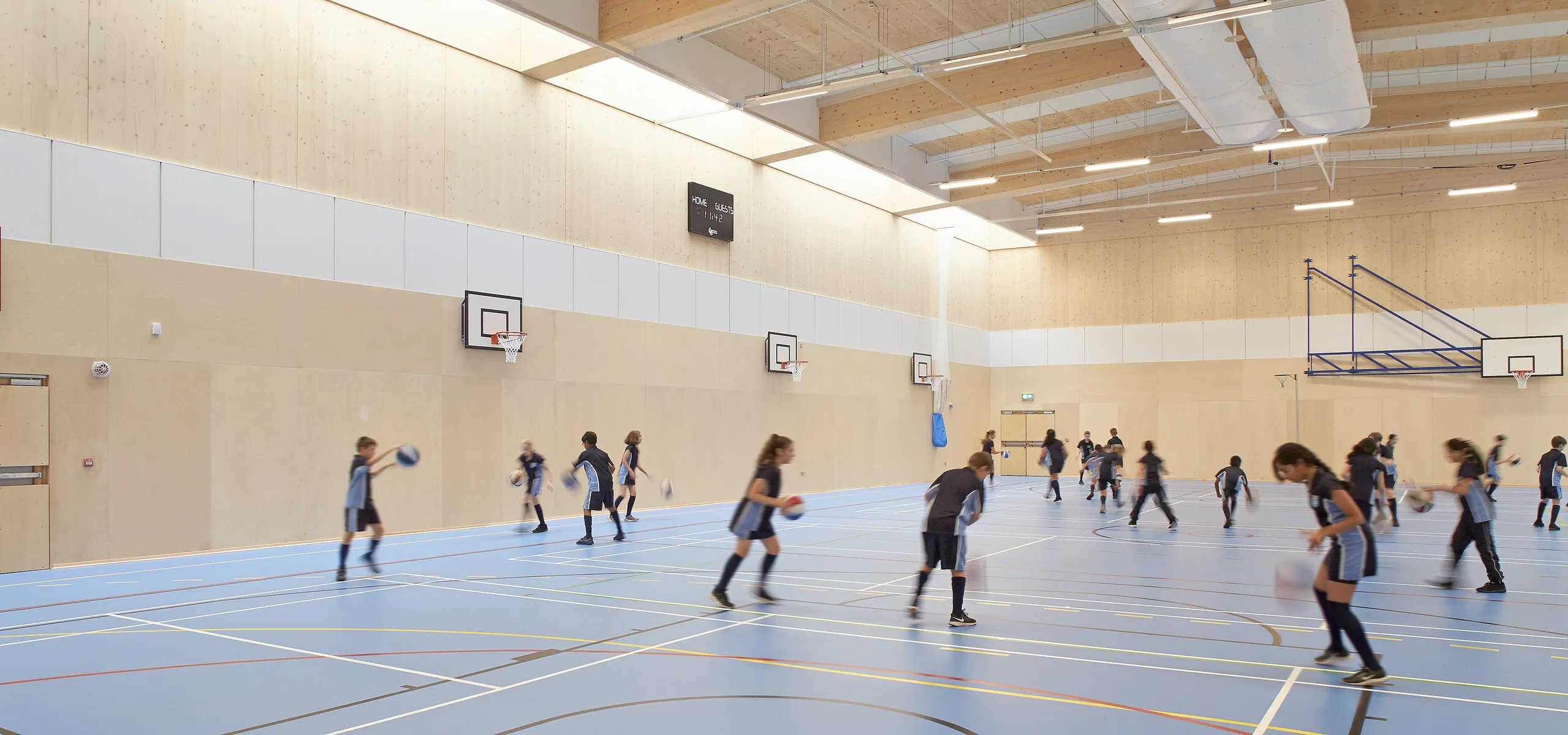 School sports hall with a timber structure, exposed beams and ventilation ducts, where students play basketball beneath suspended acoustic ceiling panels and linear lighting.