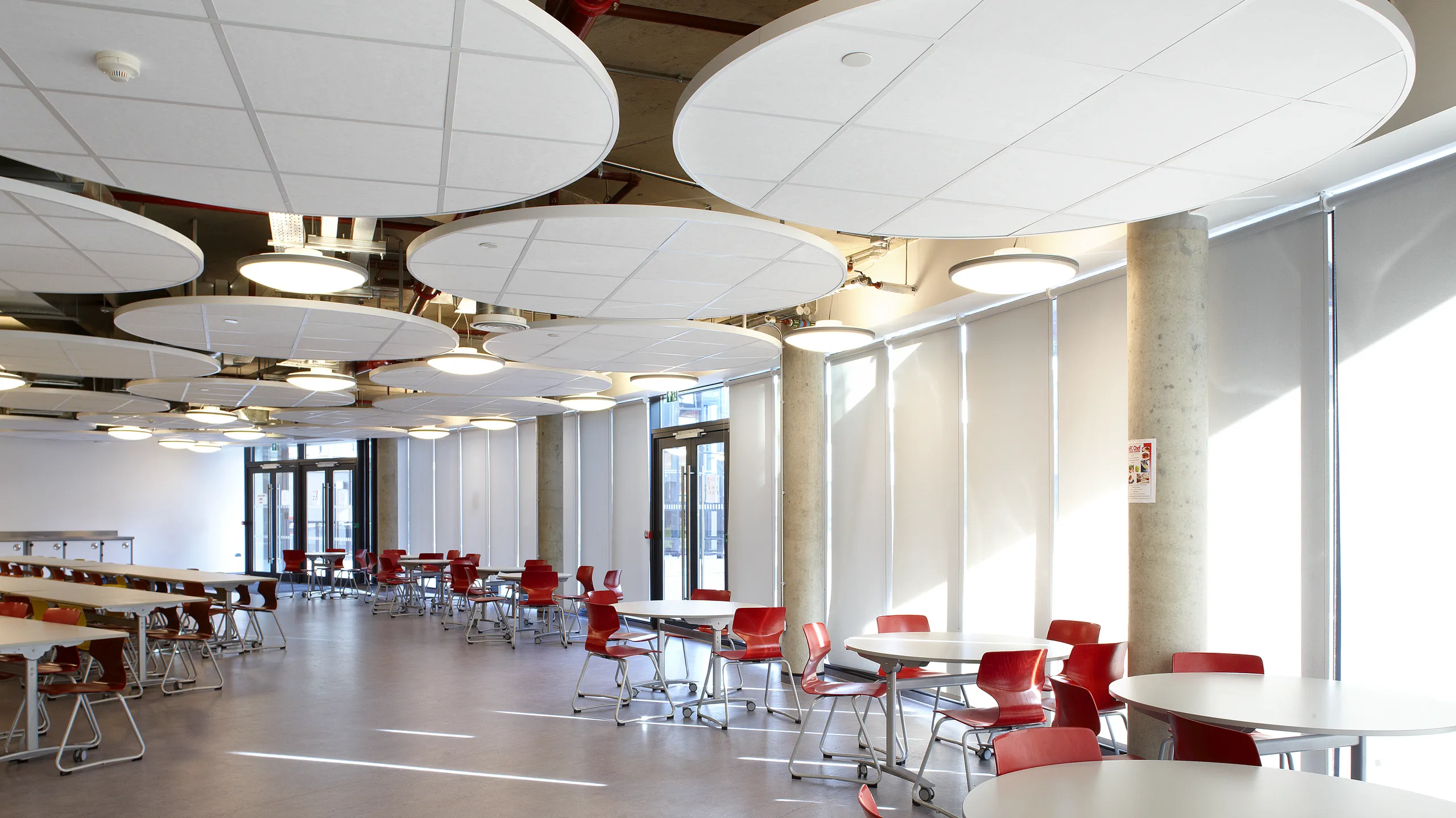 A modern dining area with a floating raft ceiling, round tables, chairs, and large circular ceiling panels. Sunlight streams in through tall windows.