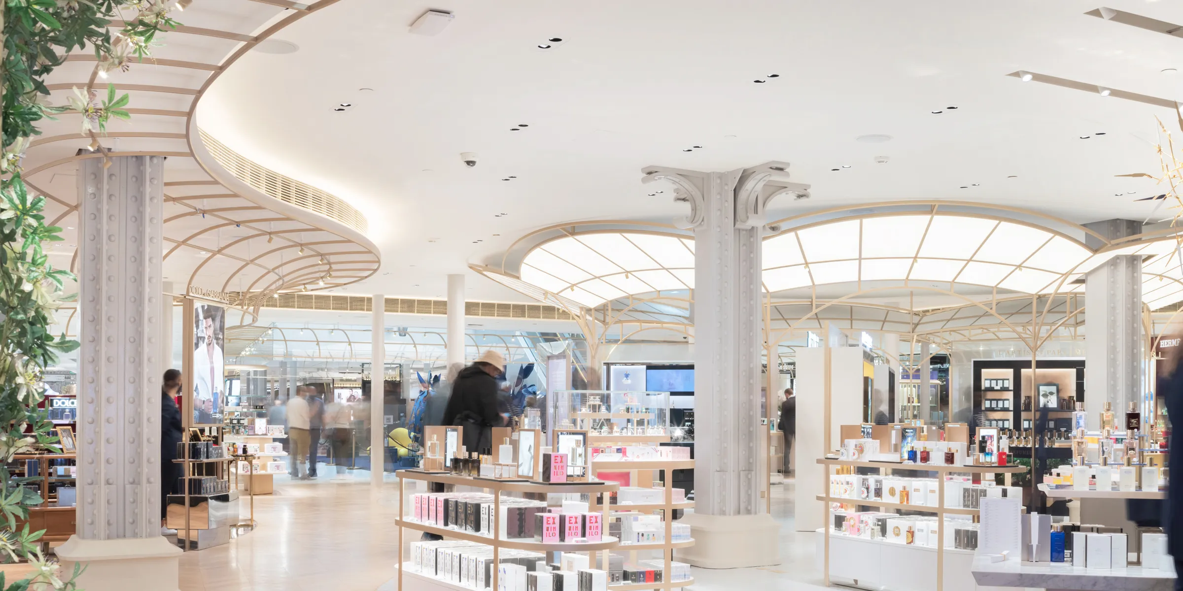 A high-end retail interior featuring a smooth seamless white acoustic ceiling, elegant lattice canopy structures, ornate columns, and island displays filled with perfumes and beauty products.