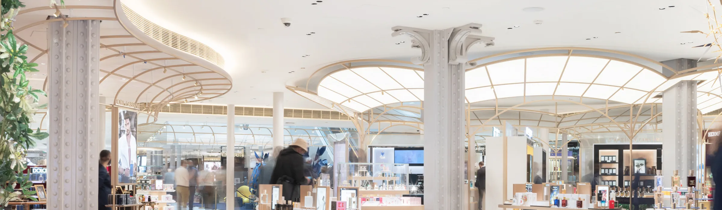 A high-end retail interior featuring a smooth seamless white acoustic ceiling, elegant lattice canopy structures, ornate columns, and island displays filled with perfumes and beauty products.
