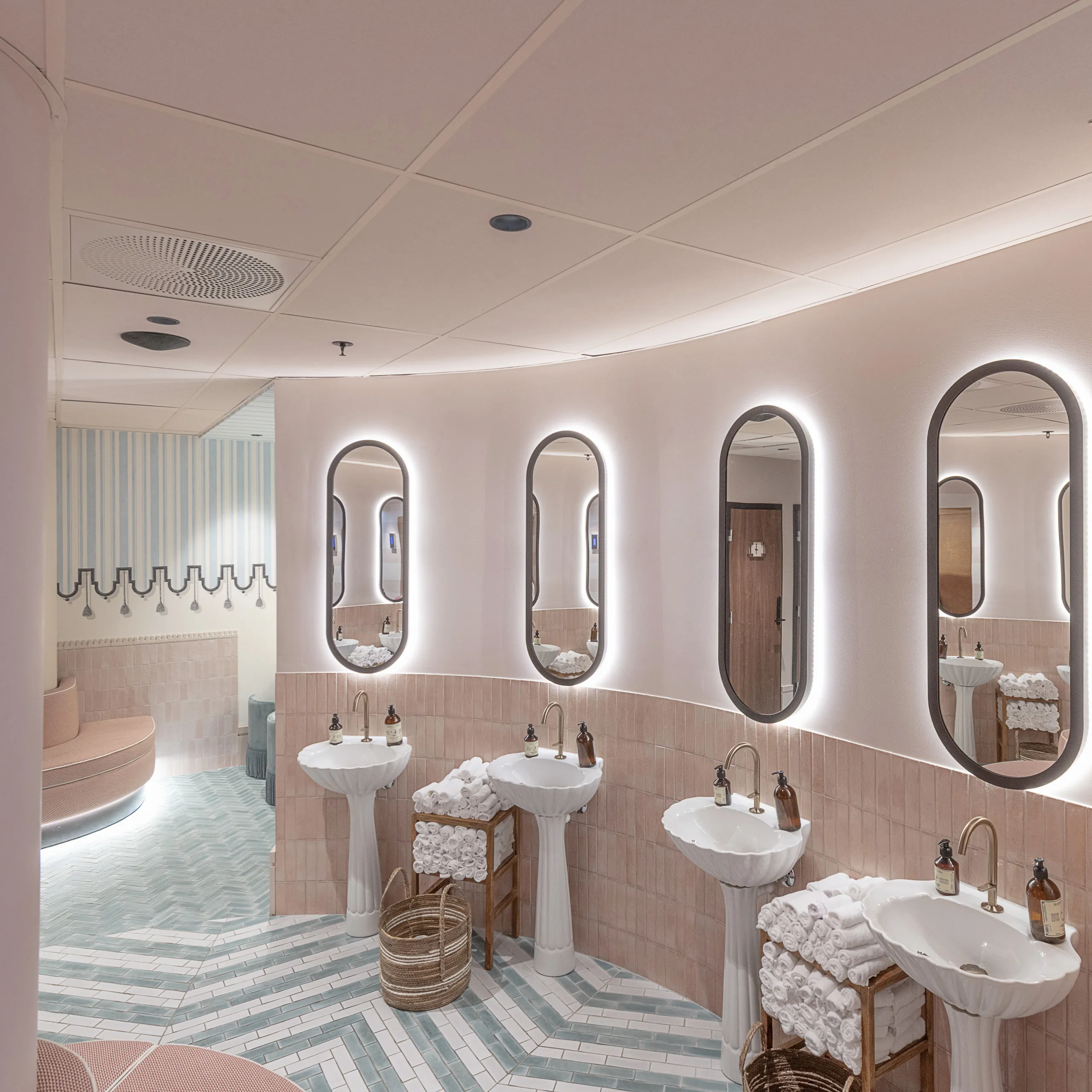 Pastel-toned restroom with curved walls, pedestal sinks, oval mirrors with backlighting, and a pastel pink acoustic tile ceiling above patterned floor tiles.