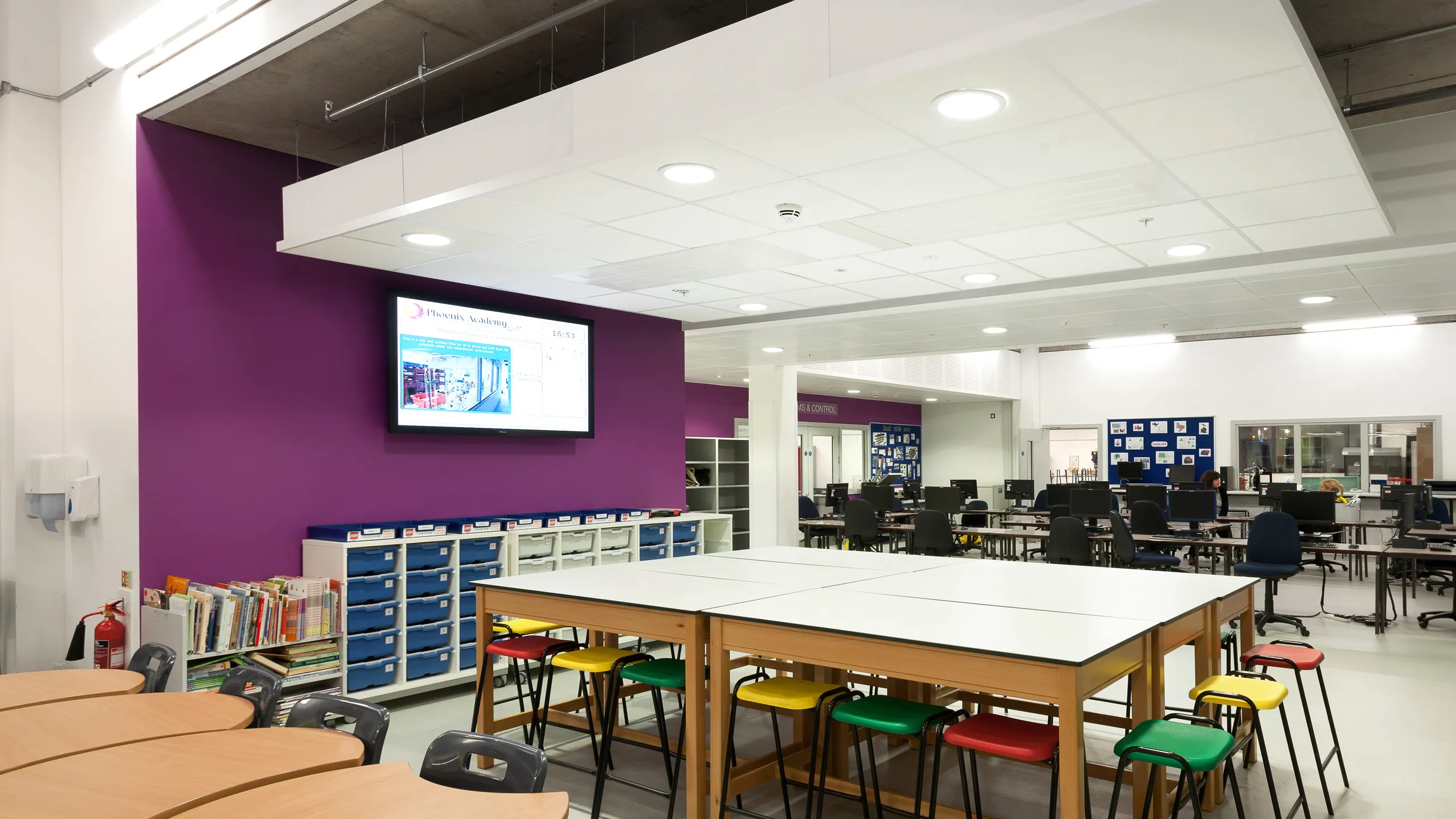 School learning space with group tables, computer workstations, and a suspended white acoustic ceiling island with integrated lighting above the classroom area.